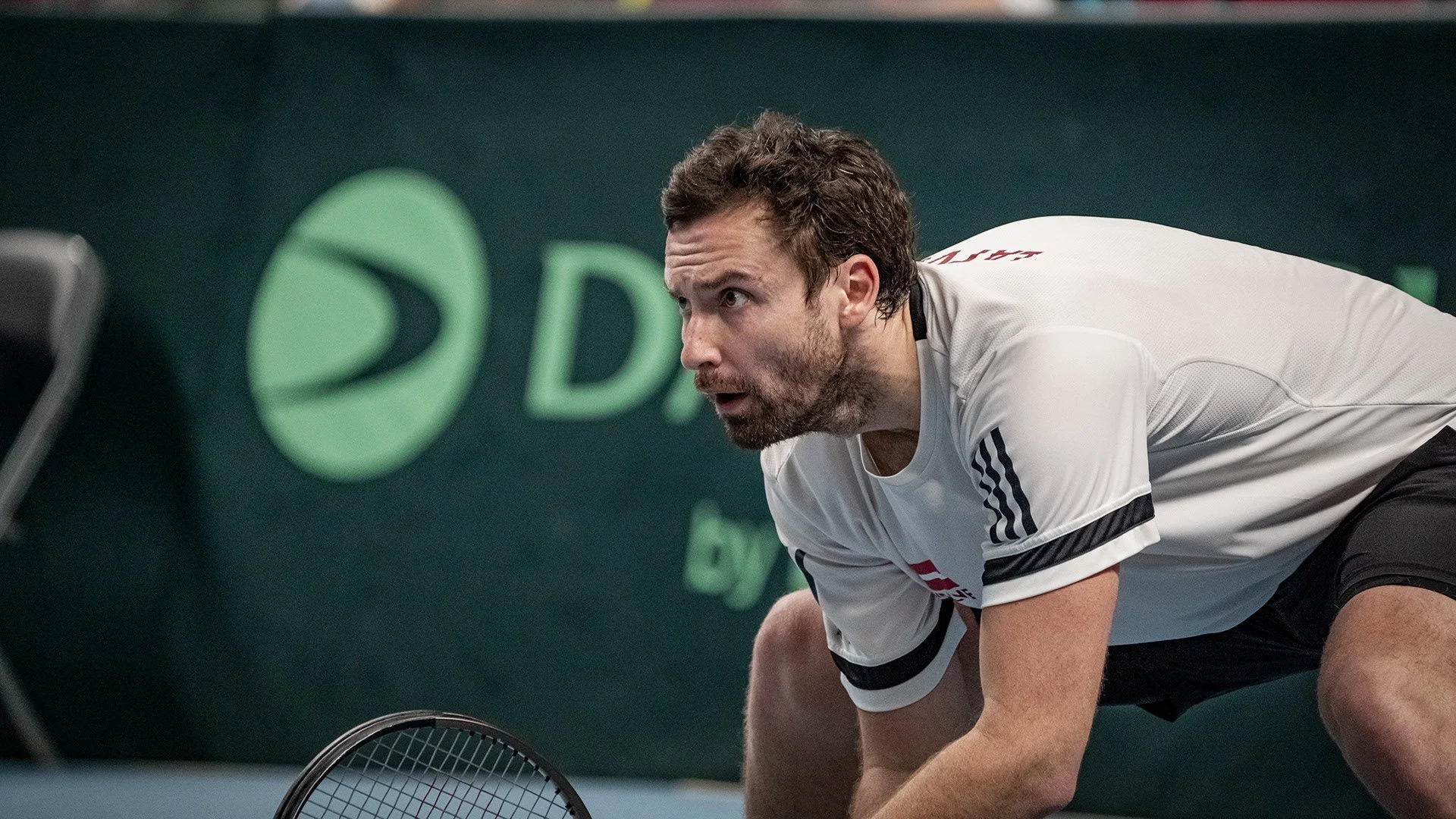 A male tennis player in a white shirt with black stripes on the sleeves, crouching on the court, holding a tennis racket, with a focused expression.