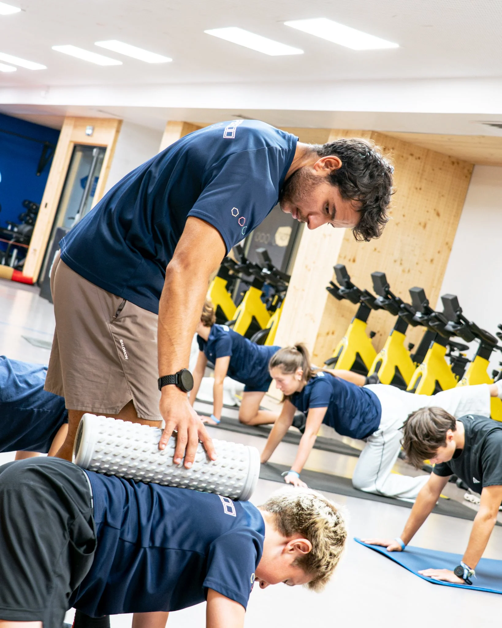 A group of people practicing yoga in a fitness studio, stretching on yoga mats with their hands on the floor and bodies bent forward. The instructor is guiding them, standing and leaning over a person in the front.