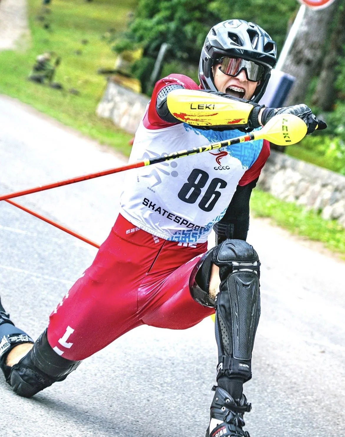 A person in protective gear and a helmet rollerblading on a paved road with greenery and stone wall in the background, holding a yellow and red baton.