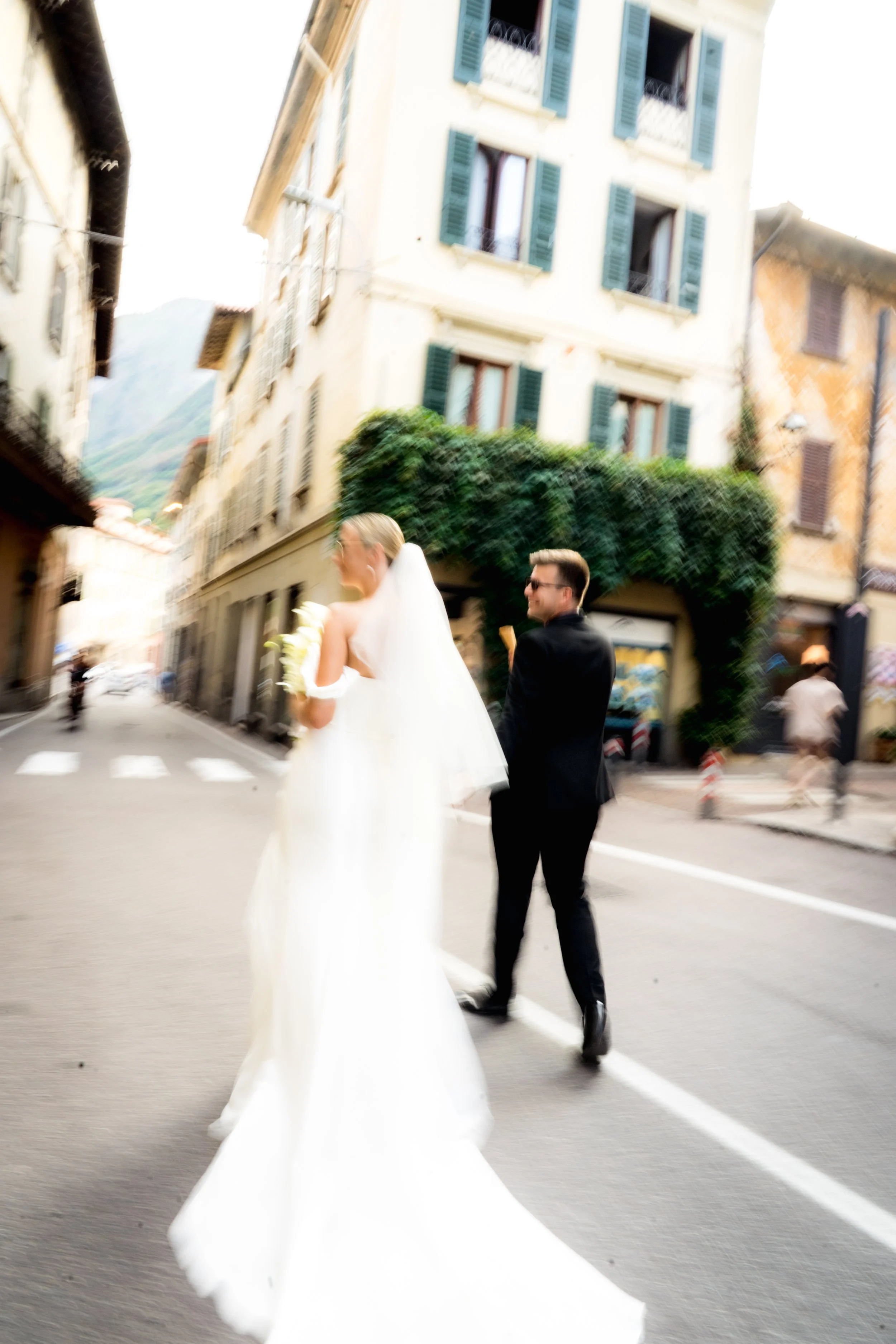 A bride and groom walking on a street in a European town with colorful buildings, windows with shutters, and greenery.