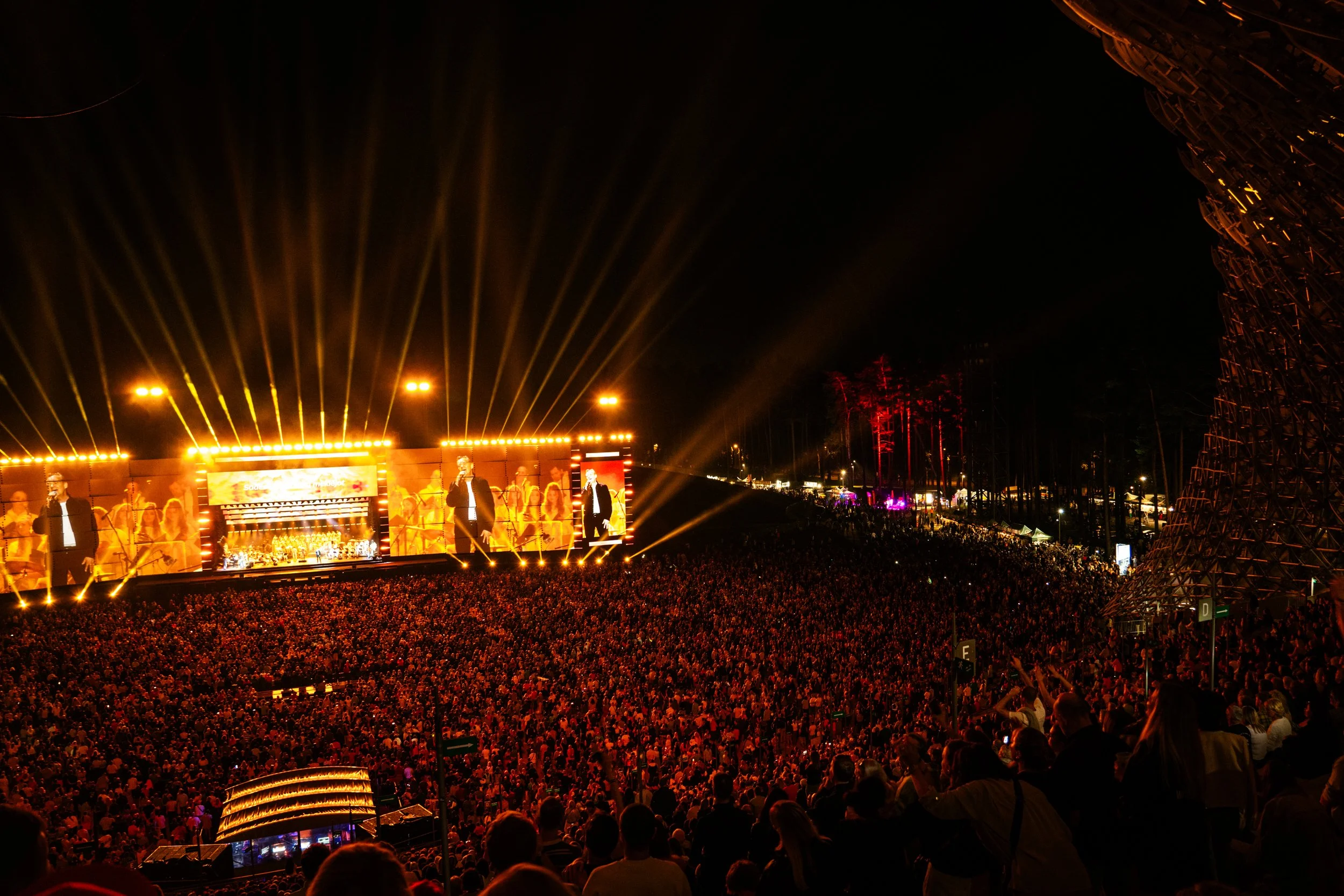 Nighttime outdoor concert with stage, performers, and a large audience, illuminated by colorful lights and laser effects.