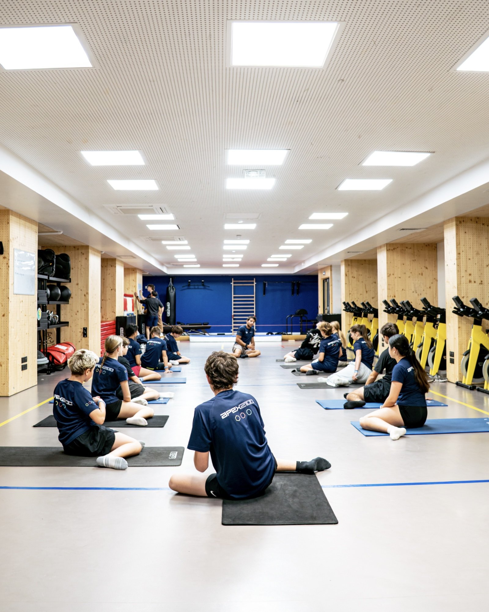 Children participating in a yoga or meditation class in a gymnasium, sitting cross-legged on mats with an instructor leading the session.