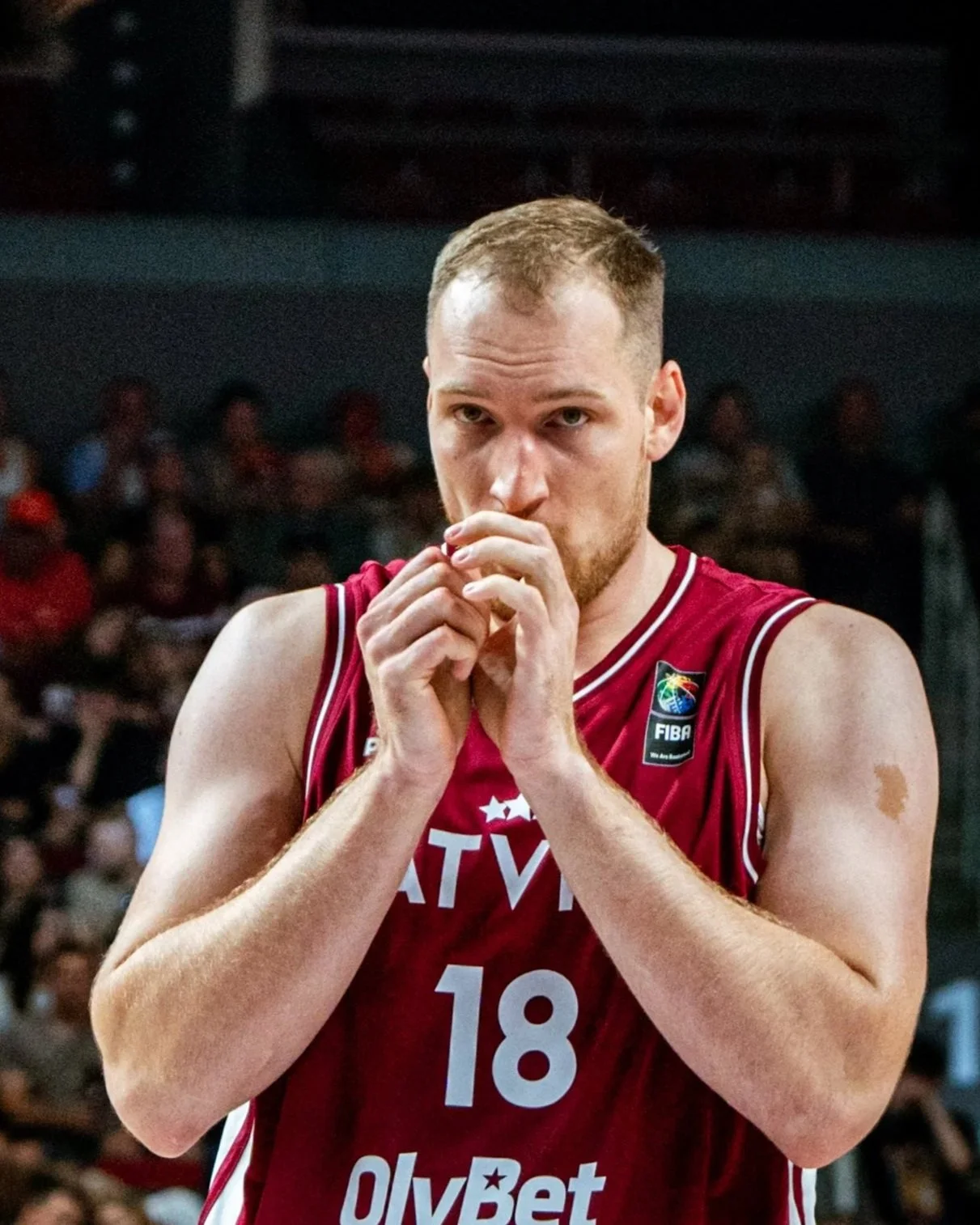 A male basketball player wearing a red jersey with the number 18 and a FIBA logo, standing on a court with a crowd in the background.