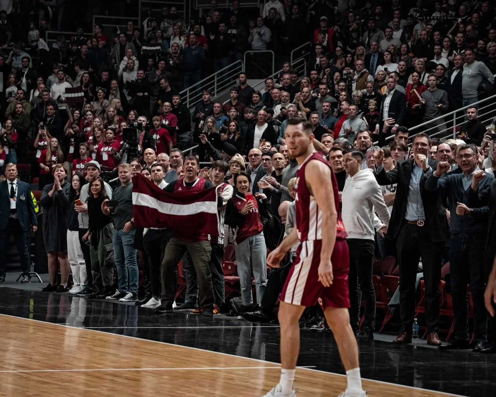A basketball player in a maroon and white uniform walking past a crowd of spectators during a game. The crowd is cheering and clapping, with some holding drinks and a flag.