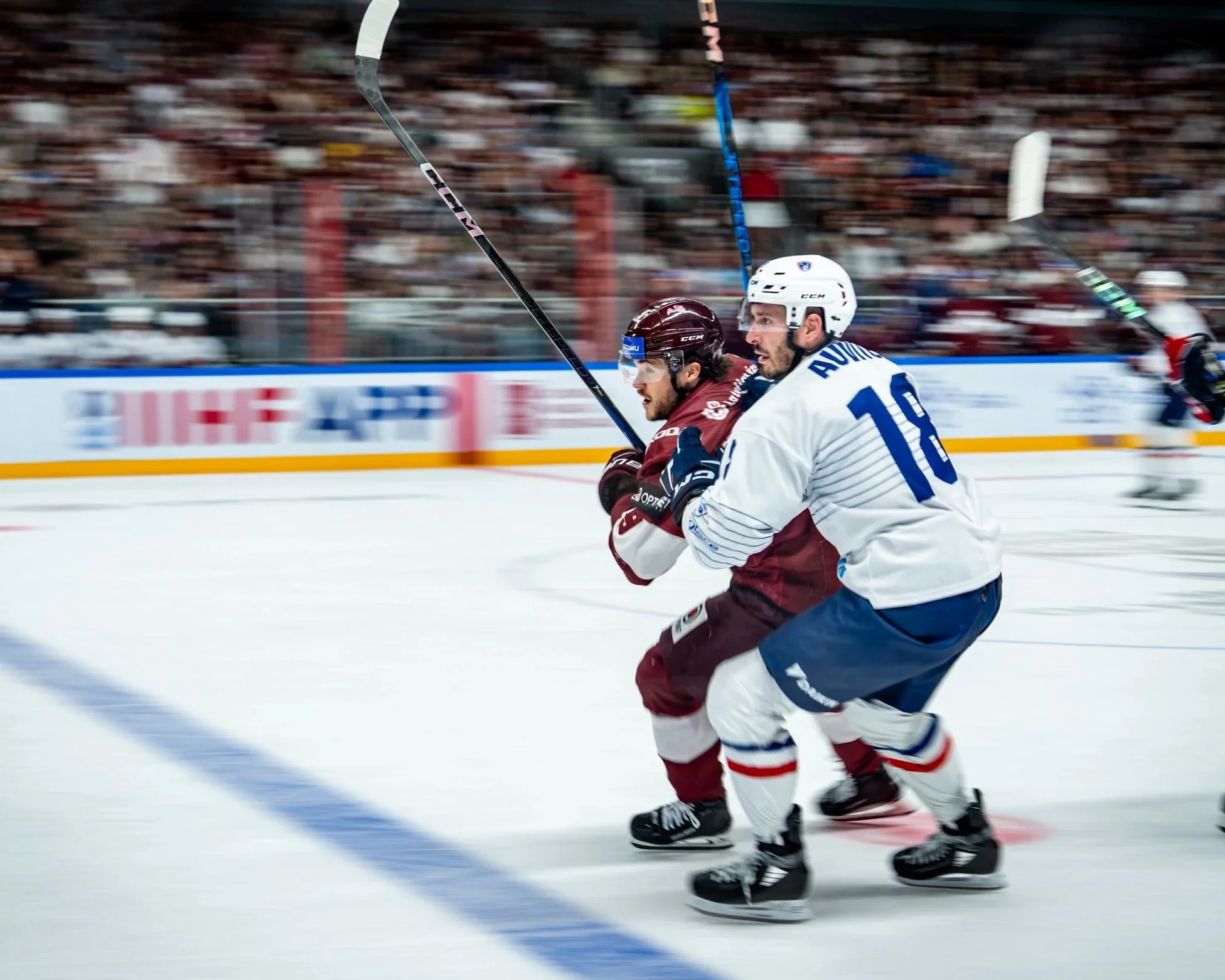 Two hockey players competing for the puck during a game, one in a white jersey and the other in a maroon jersey, on an ice rink with an audience in the background.