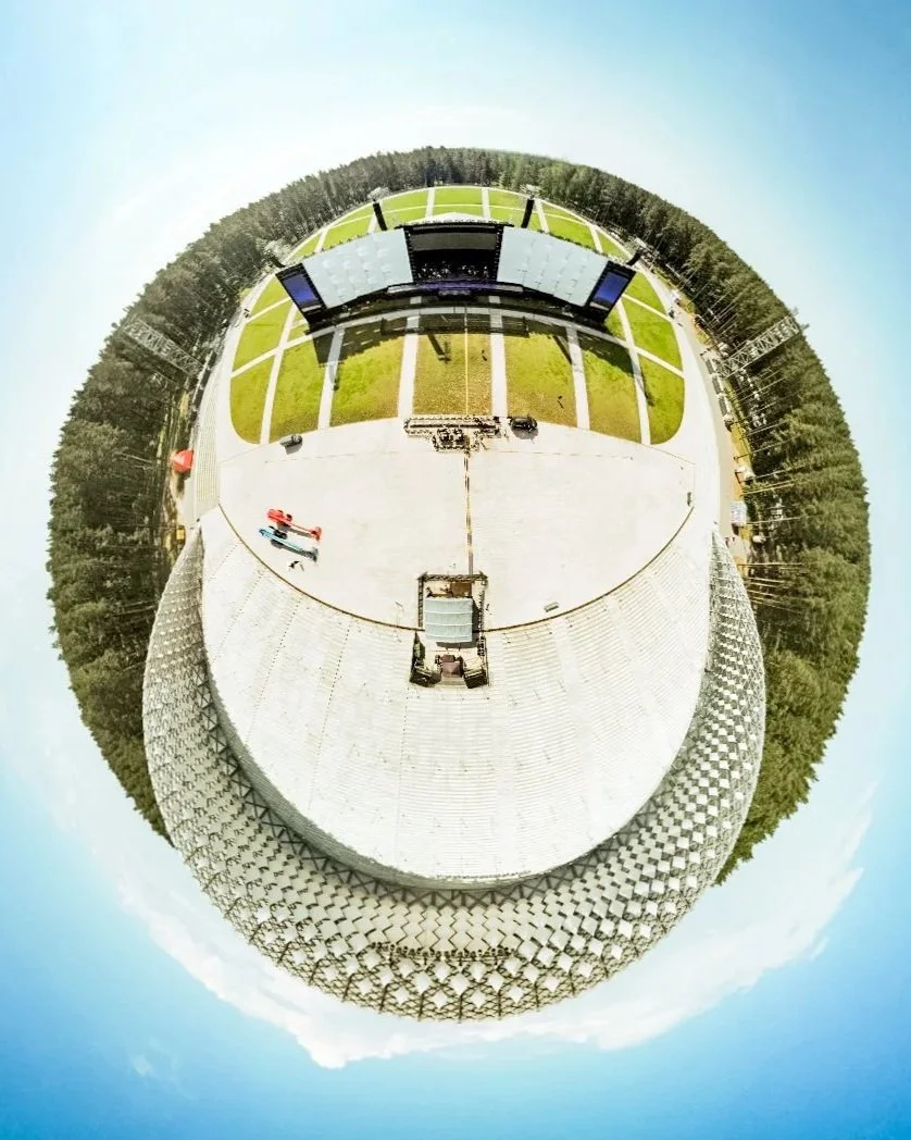 A circular fisheye view of a large outdoor stage with a screen, surrounded by grassy fields and trees, under a clear blue sky.