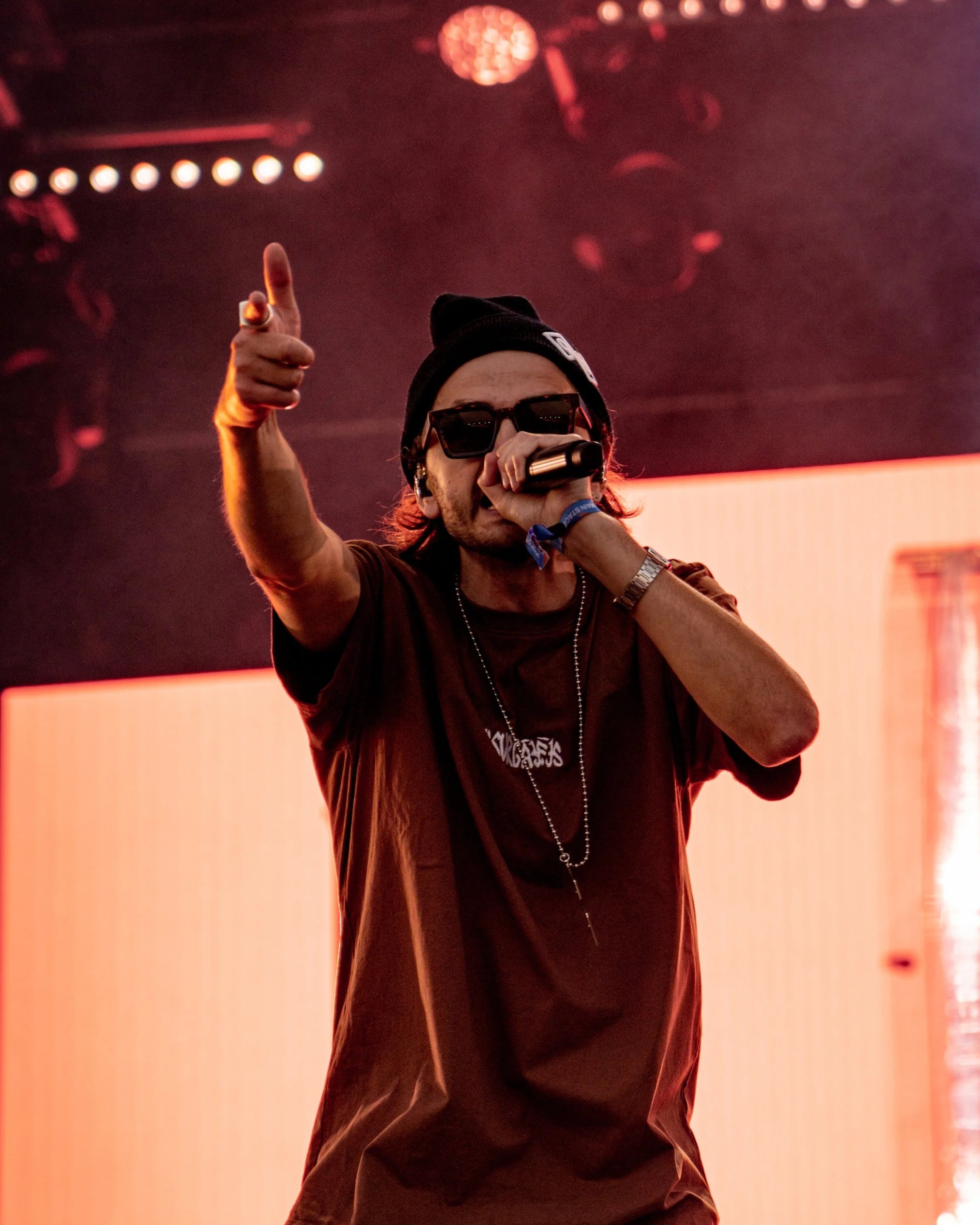 A person with long hair, wearing dark sunglasses, a black beanie, a black t-shirt, and a silver chain, holding a microphone and giving a thumbs-up during a performance on stage with a reddish-pink background and stage lighting.