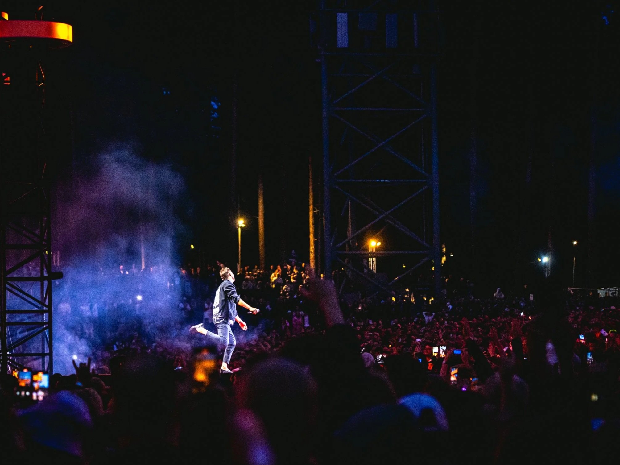 Nighttime stage performance featuring a male performer dressed in dark clothing, seemingly jumping or kneeling on stage with an audience in front, surrounded by colorful lights and smoke effects.