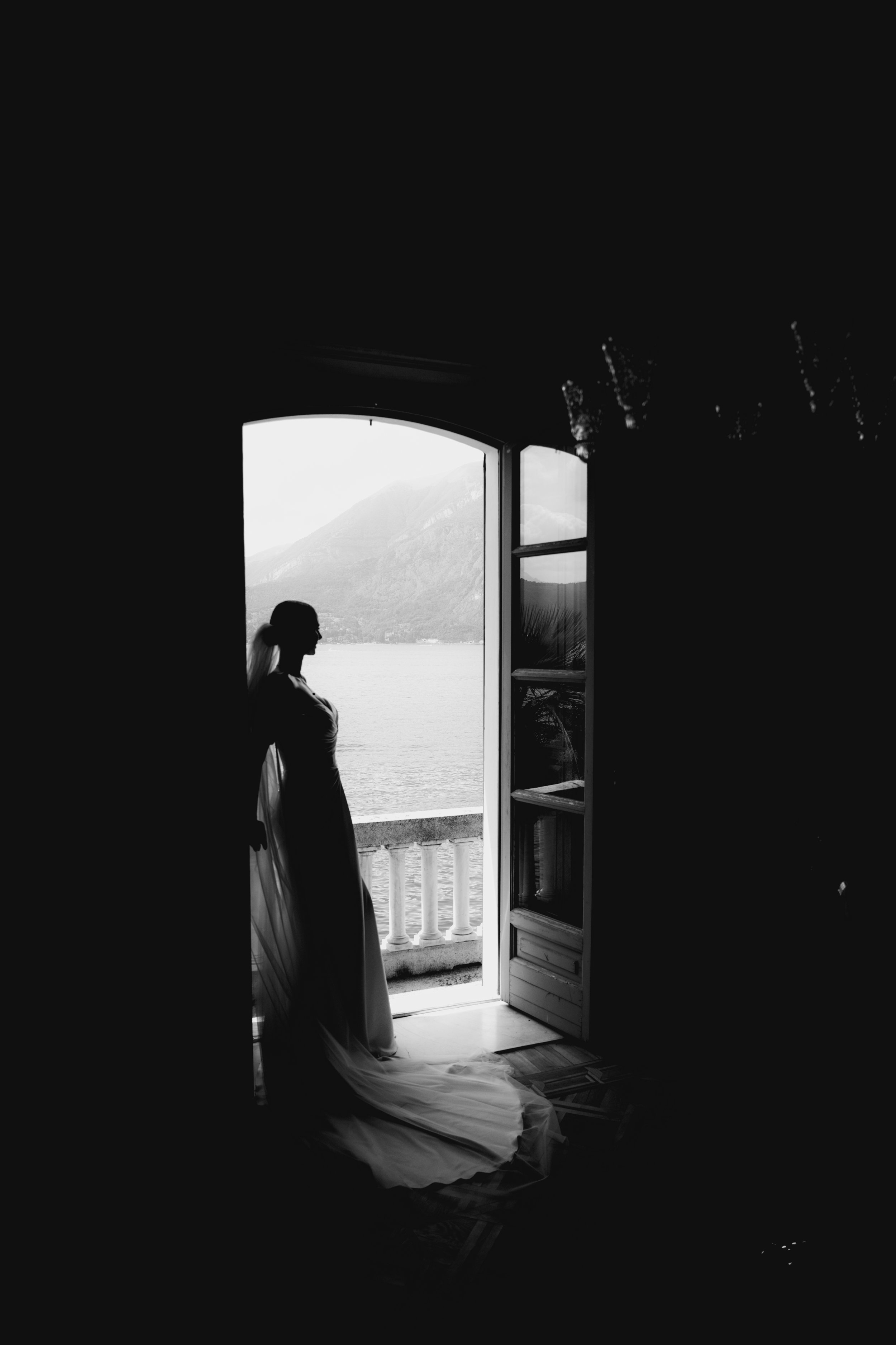 A woman in a wedding dress standing on a balcony, looking outside through an open door, with a landscape of Lake Como