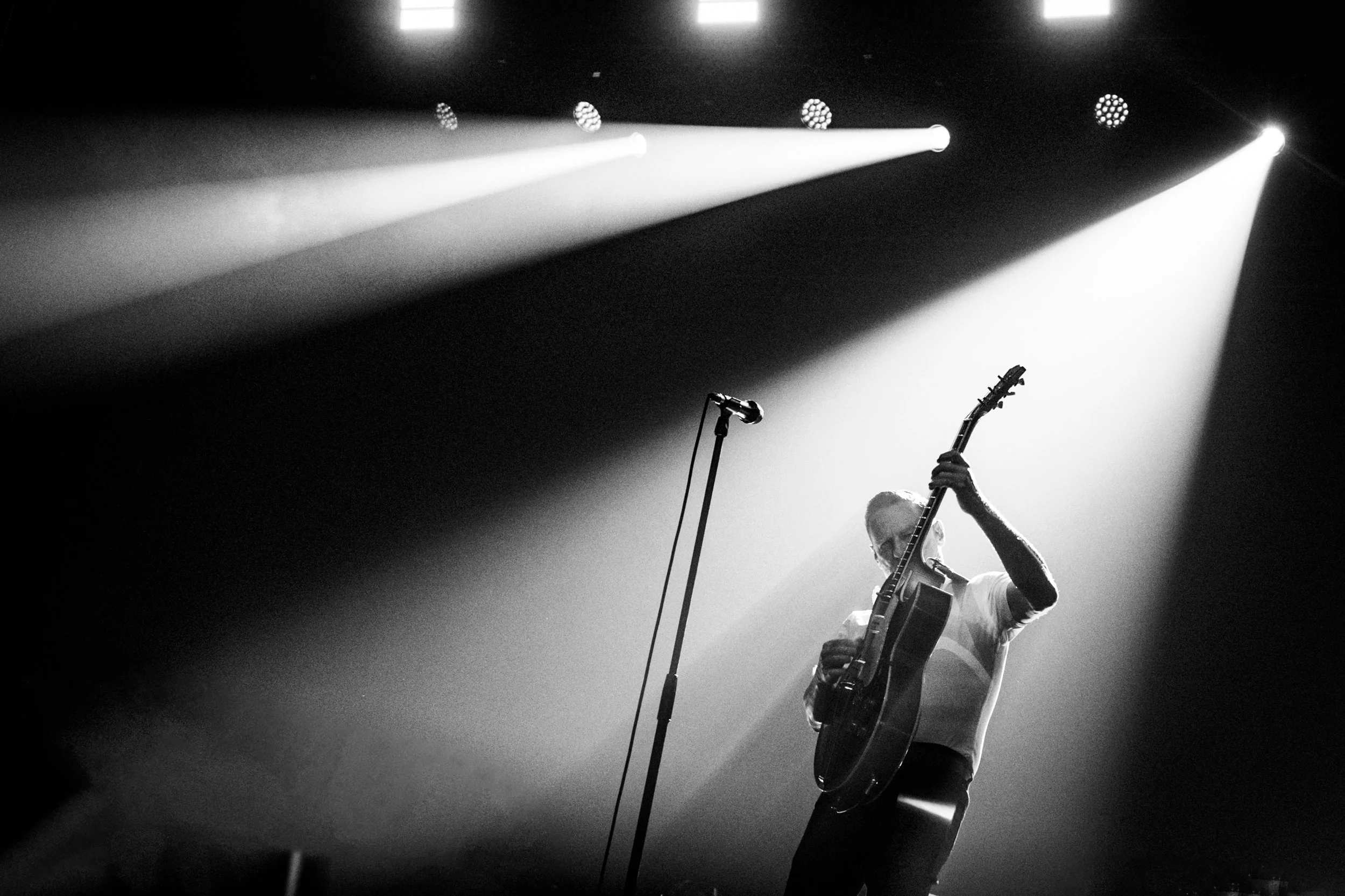 A musician playing an electric guitar on stage with bright spotlights overhead in black and white. Bryan Adams
