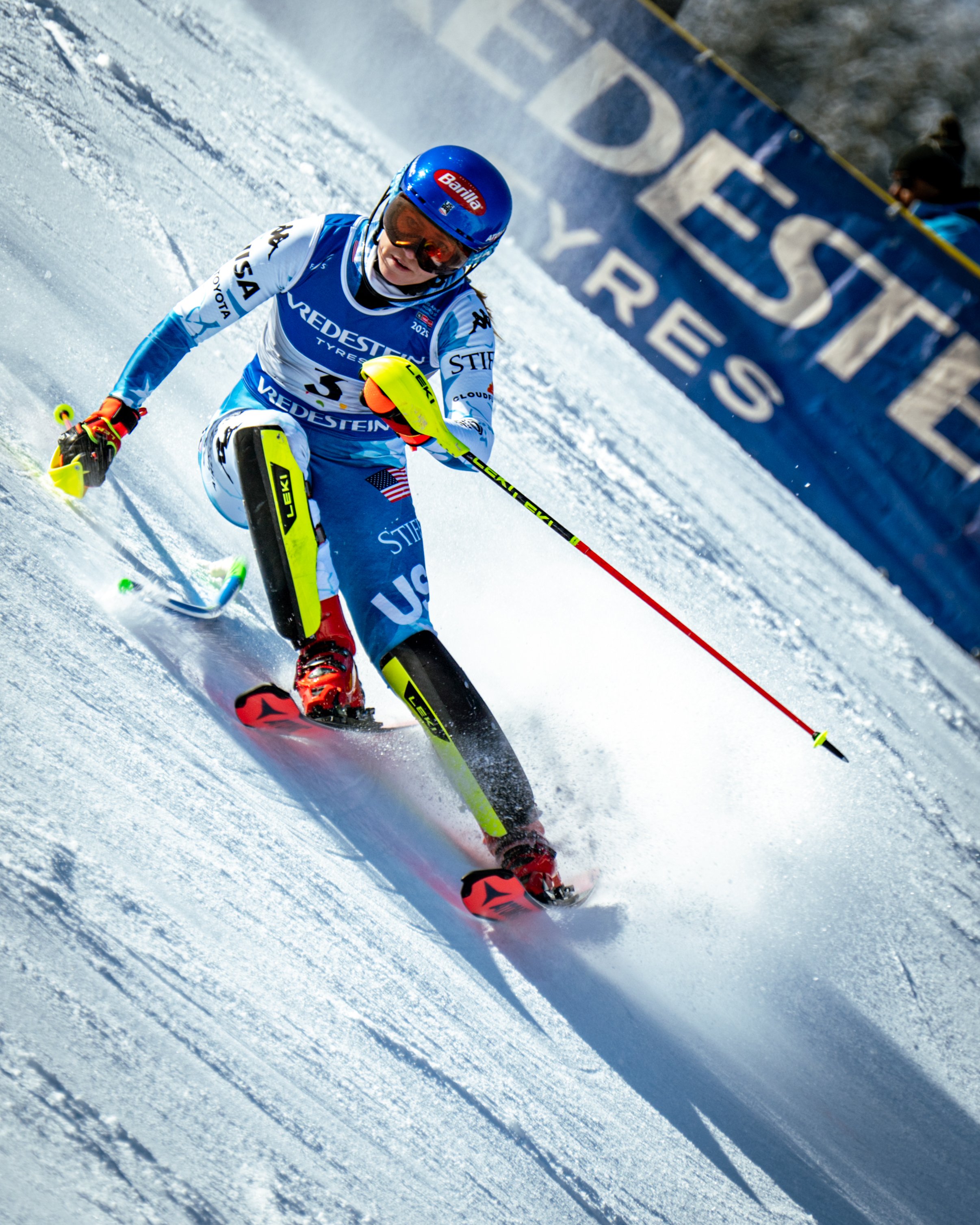 A female alpine skier in blue and white gear racing downhill on snow, wearing a blue helmet and goggles, with a banner and snow in the background.