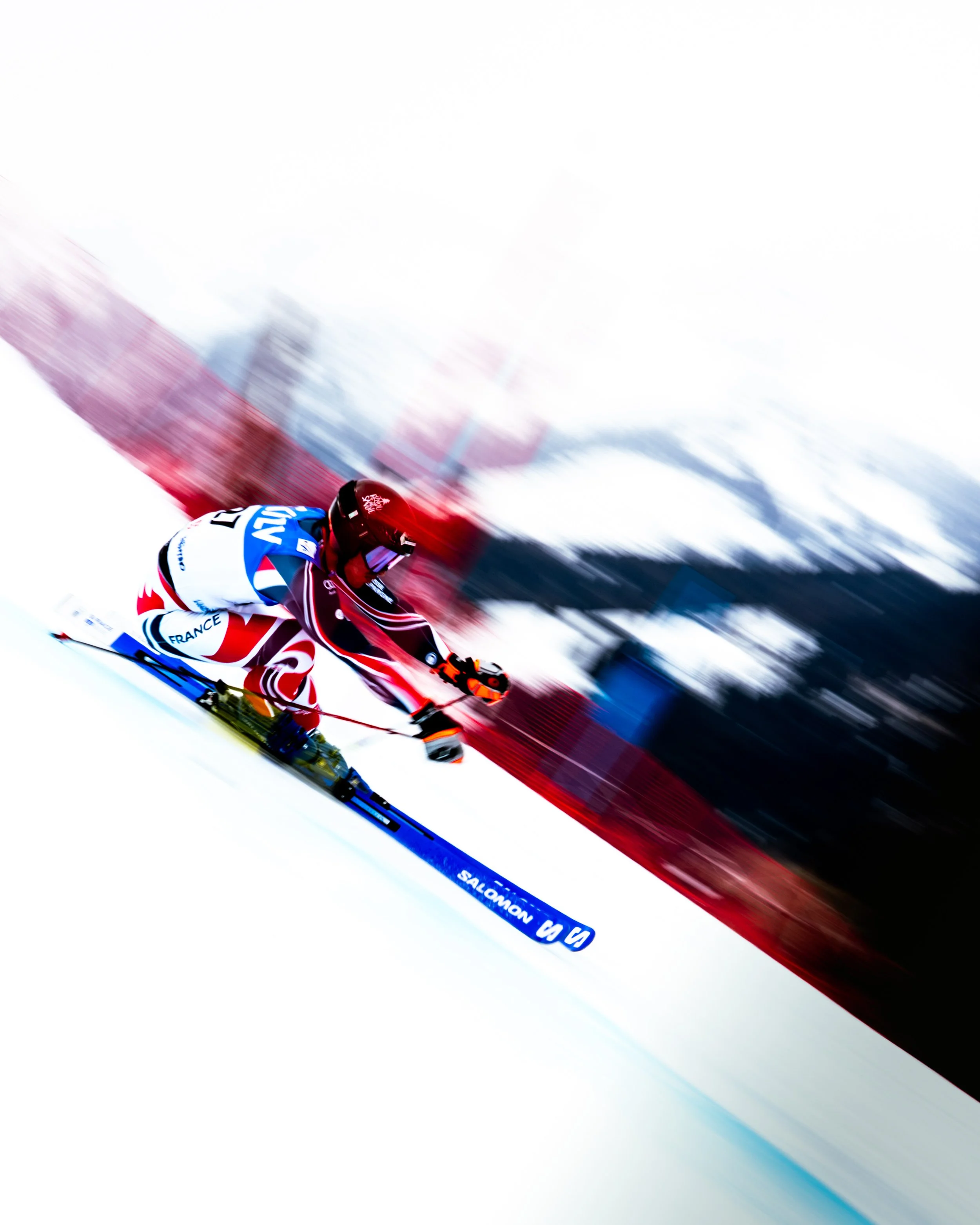A skier in a red, white, and blue suit with French flags, wearing a red helmet, speeds down a snow-covered slope as the photo is taken at an angle, capturing motion blur in the background.