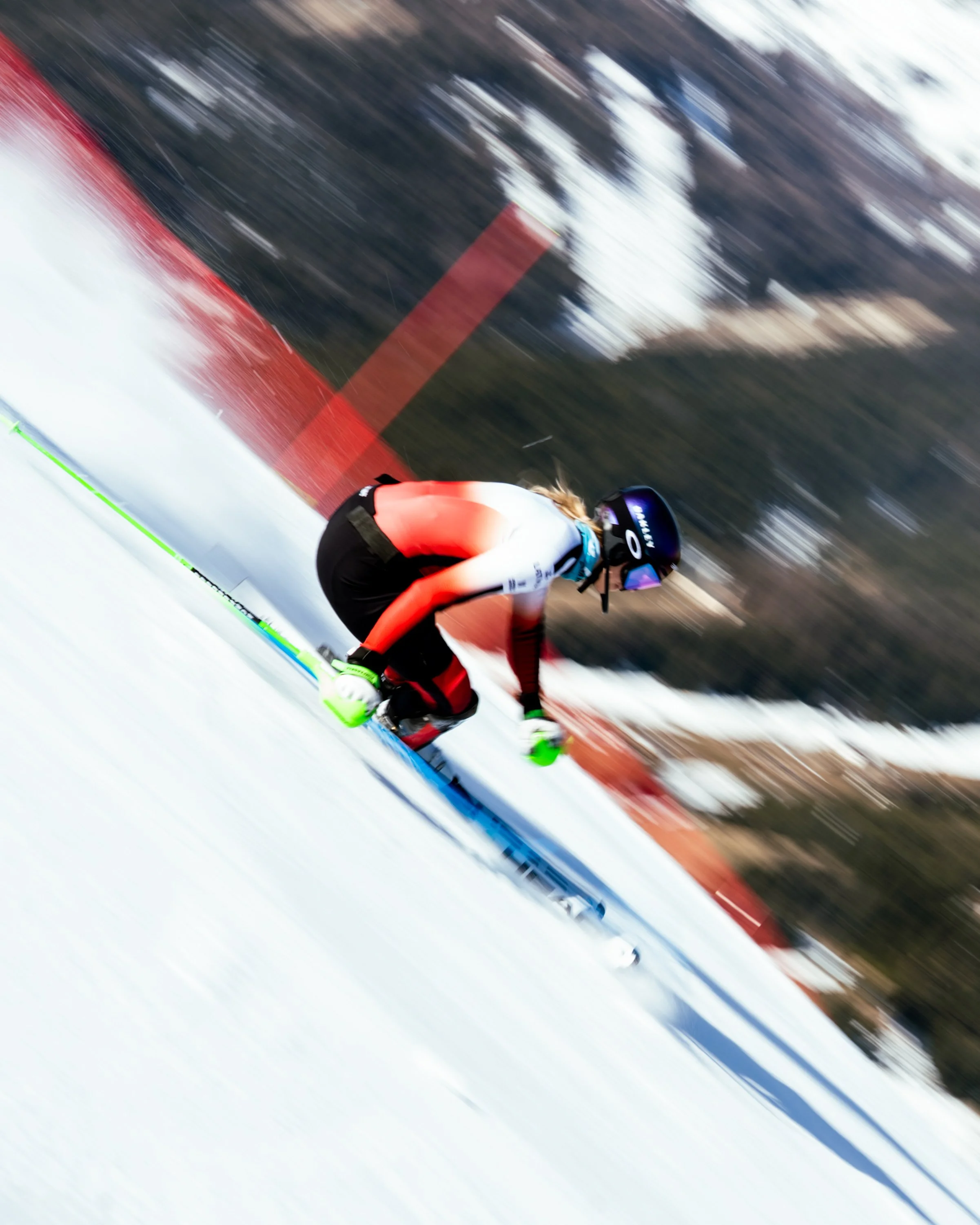 A skier in a race leaning forward on snow, with a background showing a snow-covered landscape and trees, captured with motion blur.