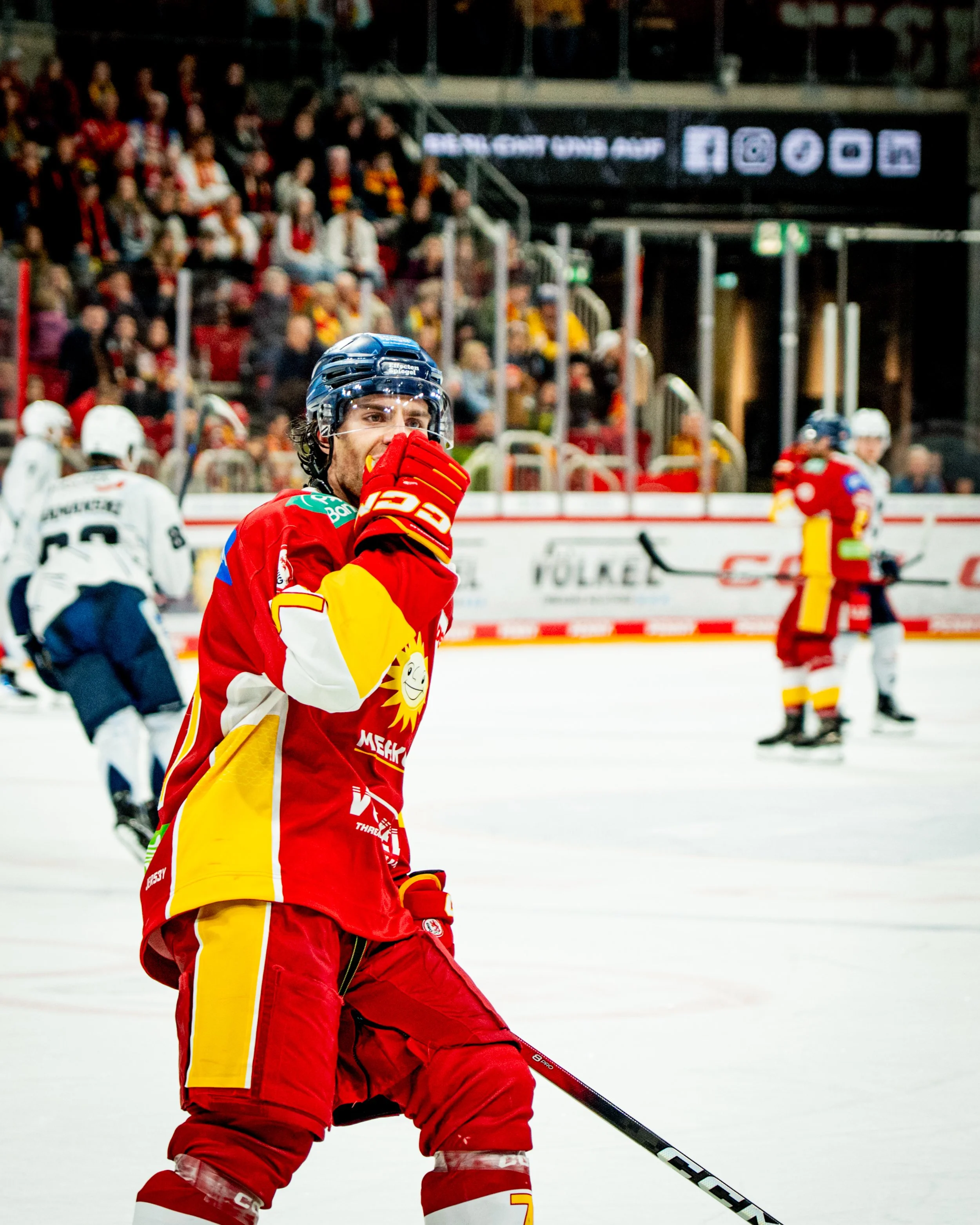 A hockey player in a red and yellow uniform with a blue helmet, holding a hockey stick, on the ice rink during a game with other players and spectators in the background. Düsseldorfer EG