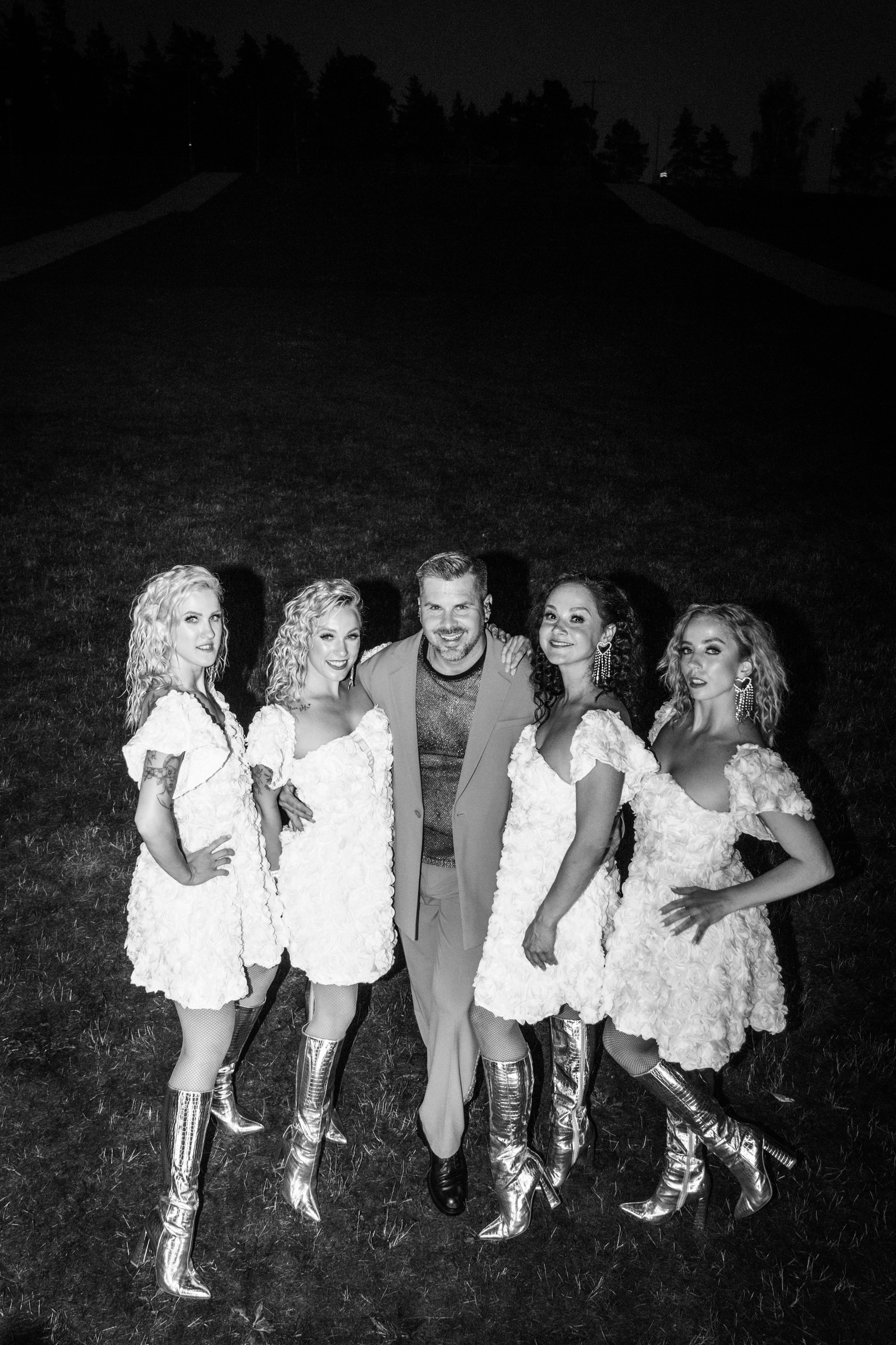 A group of five women in matching white lace dresses and knee-high shiny silver boots standing outdoors at night, with a man in a blazer and patterned shirt in the center, all smiling and posing for the camera on a grassy area.