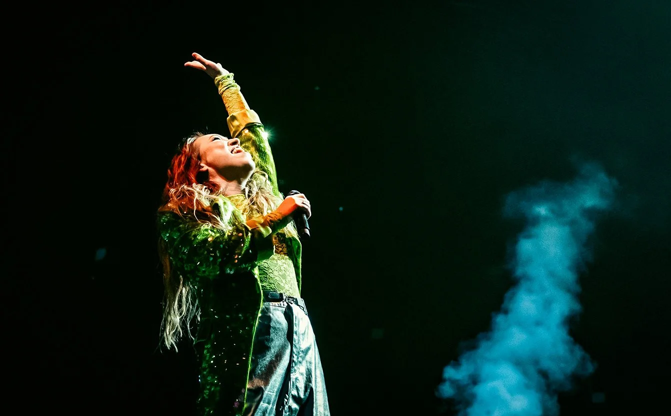 A singer performing on stage, holding a microphone in her right hand, with her left arm raised and head tilted back, smiling under green stage lighting, with blue smoke in the background.