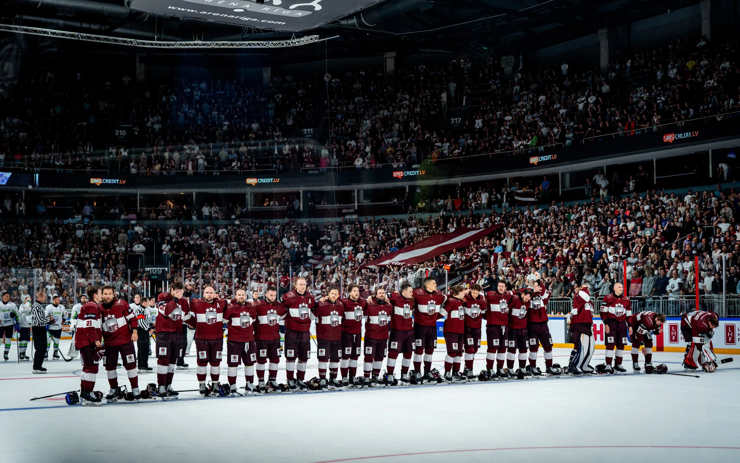 Ice hockey players in maroon jerseys standing on the ice rink with their helmets on, facing toward the audience, during a professional game. The arena is filled with spectators in the stands, and the players are lined up as a team before a game or du