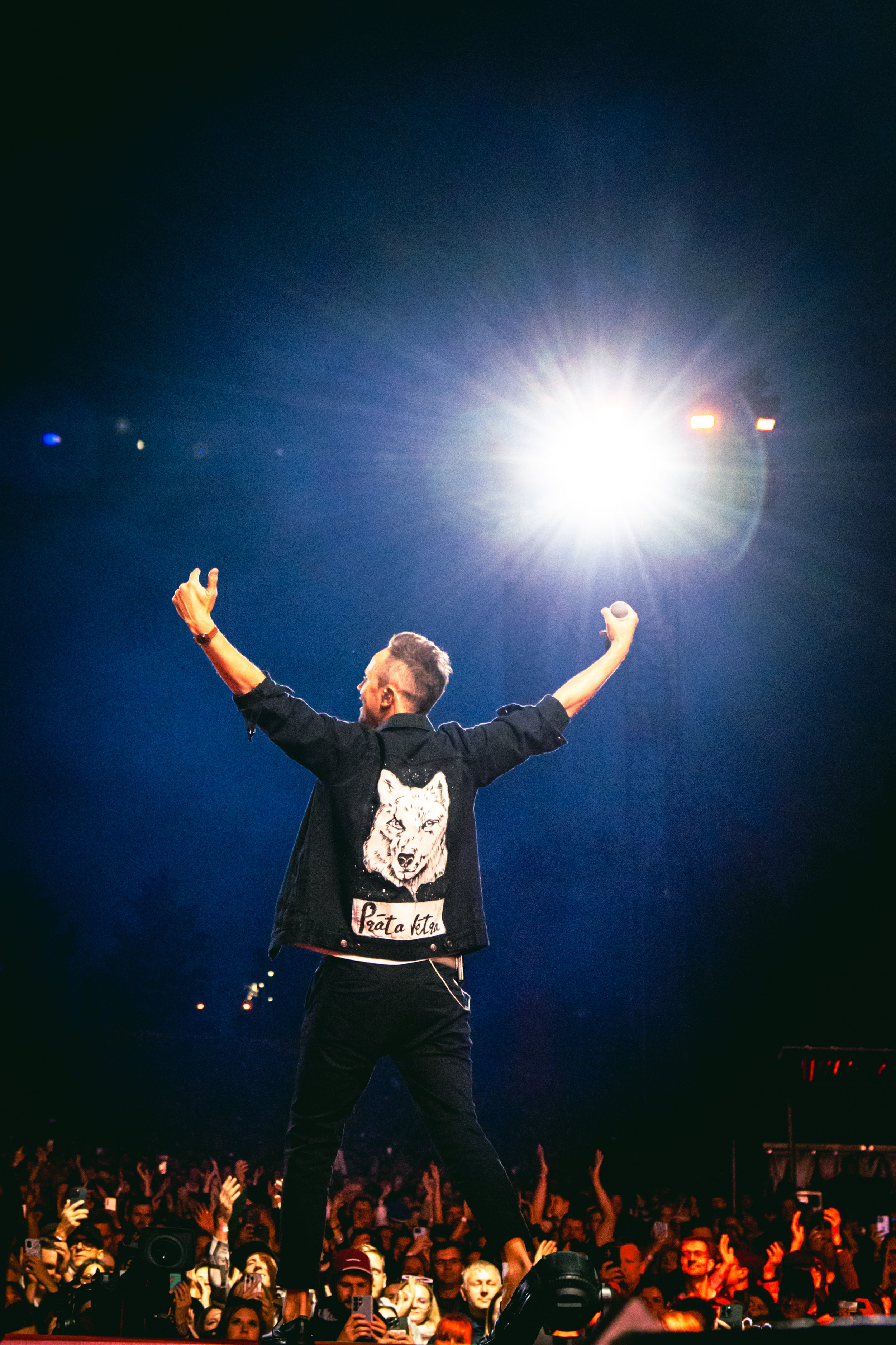 Performer on stage at a concert, arms raised, facing a large crowd, with bright stage lights overhead, audience members holding up phones. Prāta vētra