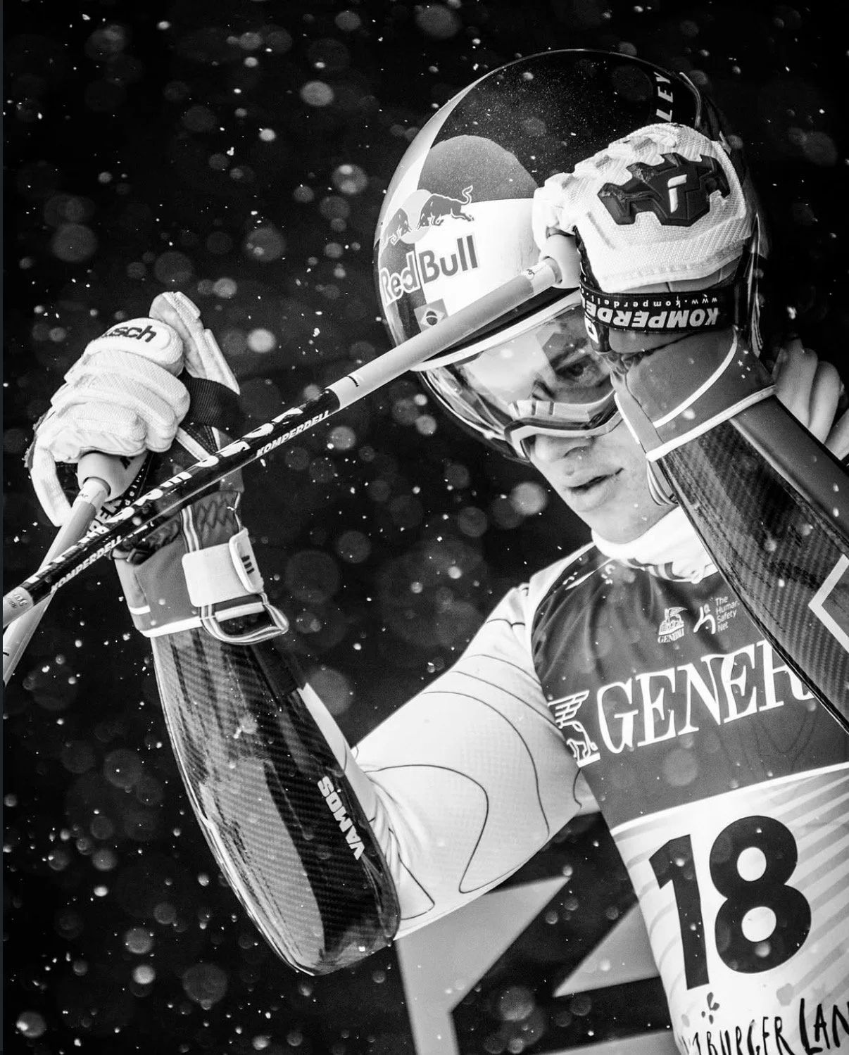 A skier wearing a helmet with a Red Bull logo, goggles, gloves, and a jersey numbered 18, adjusting their ski poles in a snowy environment.