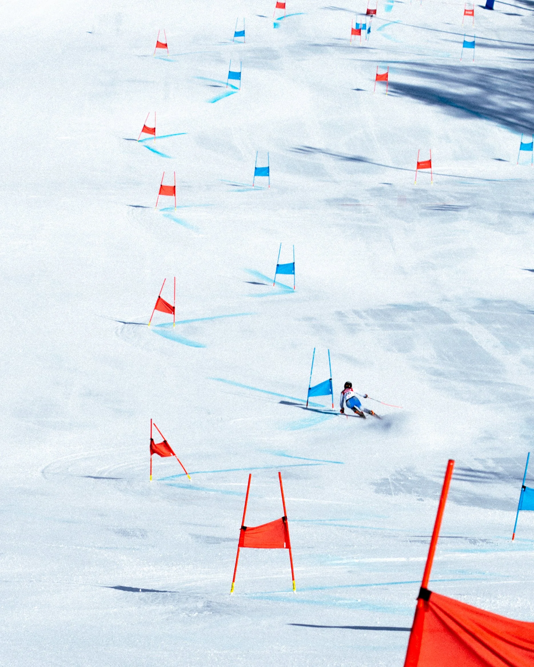 A skier navigating a slalom course on a snowy slope, passing red and blue gates with sunlight casting shadows.