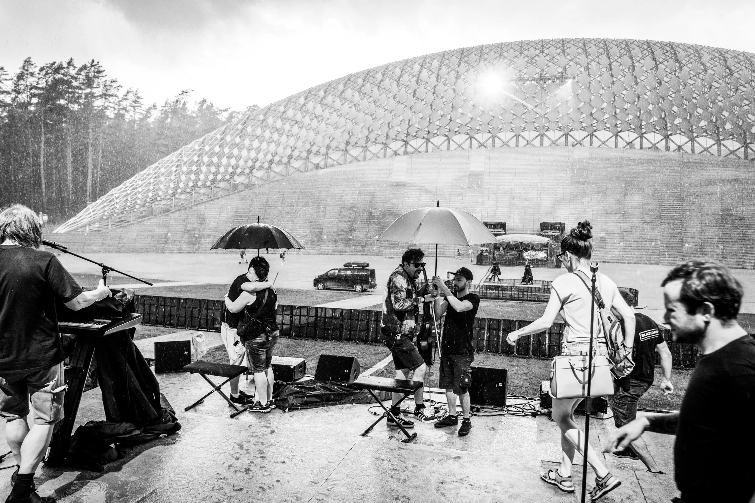 People gathered outdoors during a rain shower with umbrellas, near a modern architectural building, some playing musical instruments, others socializing, in black and white. LIELKONCERTS