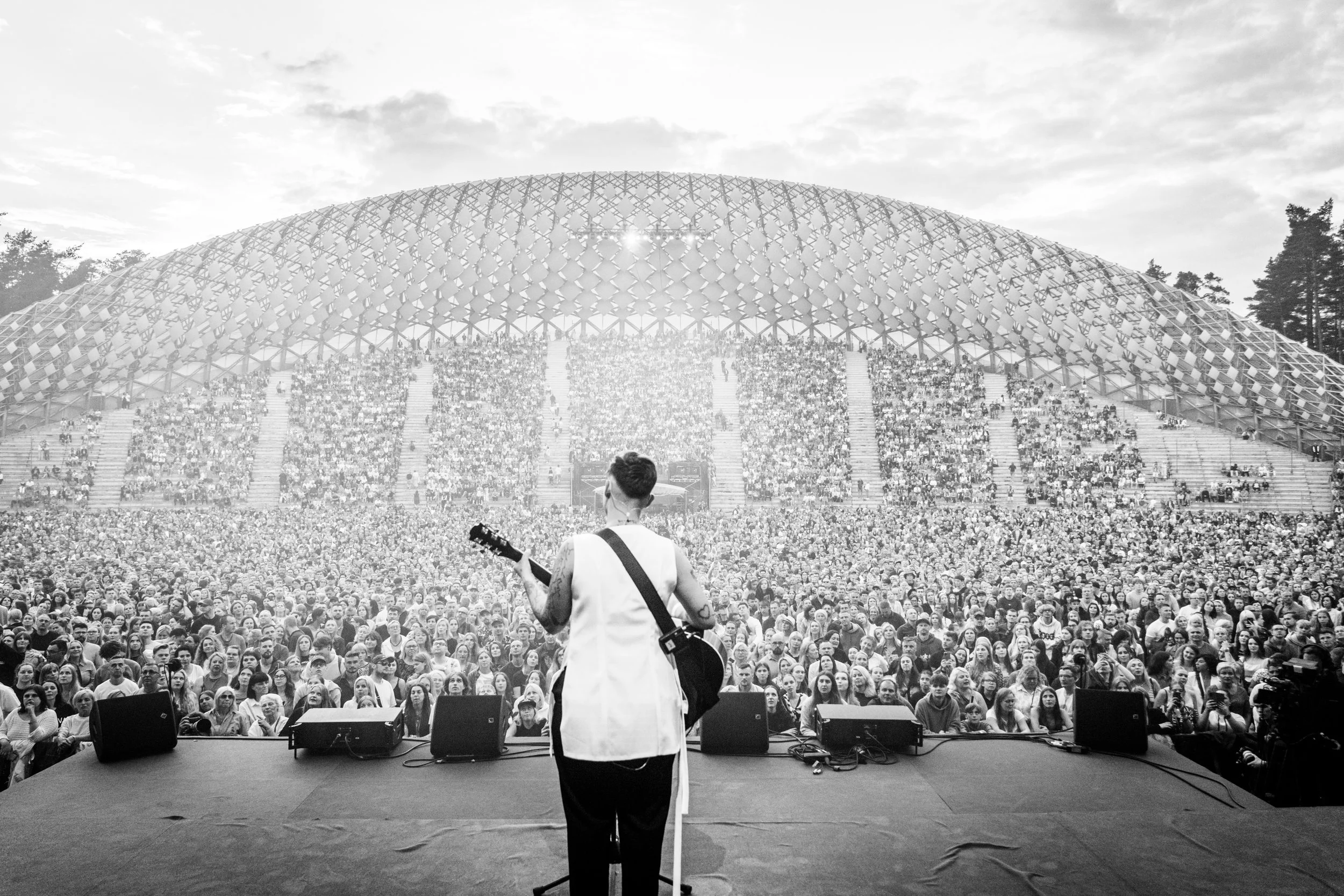 A musician on stage playing guitar in front of a large crowd at an outdoor concert during the daytime.
