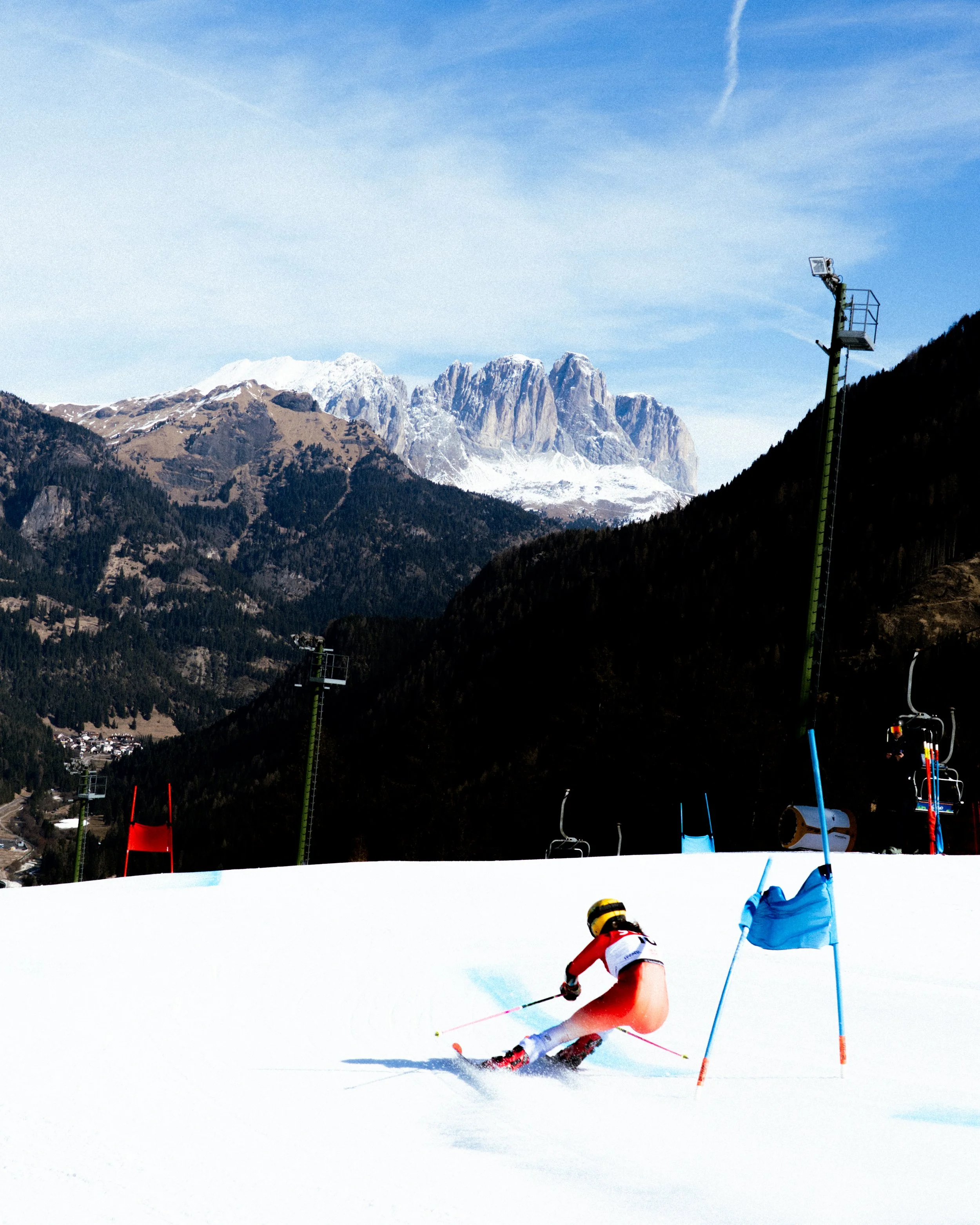 A skier in a red and white suit skiing downhill on snow with poles, wearing a helmet and yellow goggles, surrounded by a snow-covered ski slope, with ski equipment and flags, mountains in the background, and ski lift towers visible.