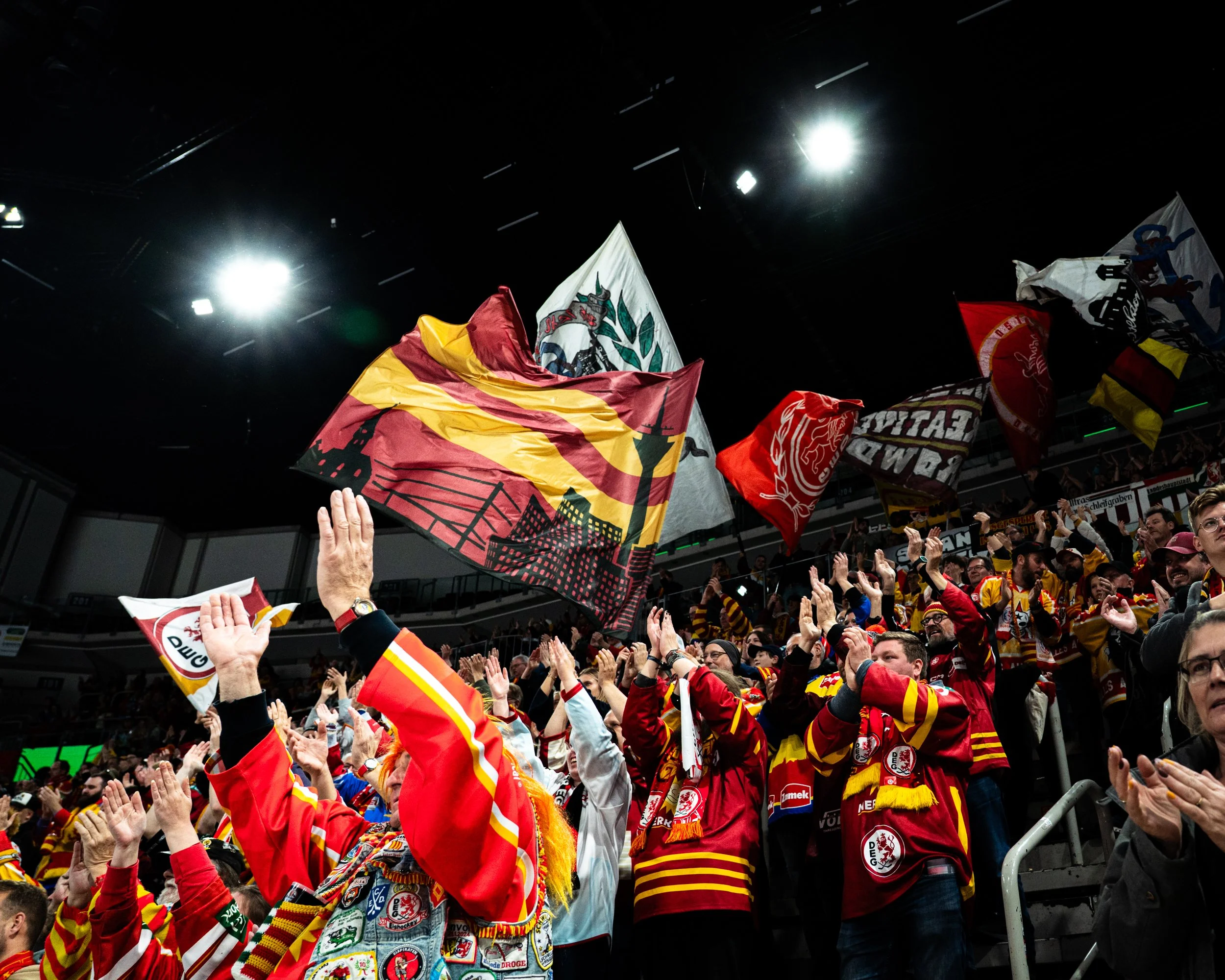 Crowd of sports fans wearing red and yellow jerseys, waving flags and cheering inside an arena with bright overhead lights.
