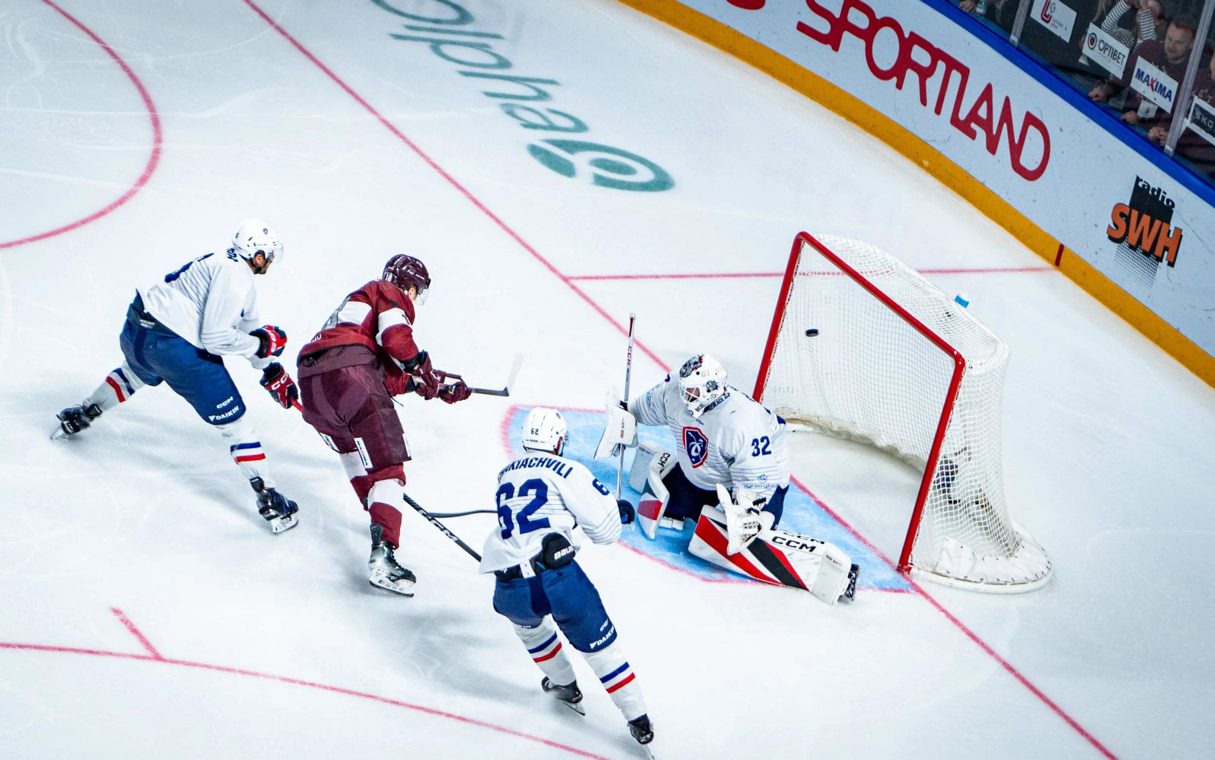 Ice hockey players near the goal, one attempting to score, while the goalie and defenders try to block the shot on the ice rink.
