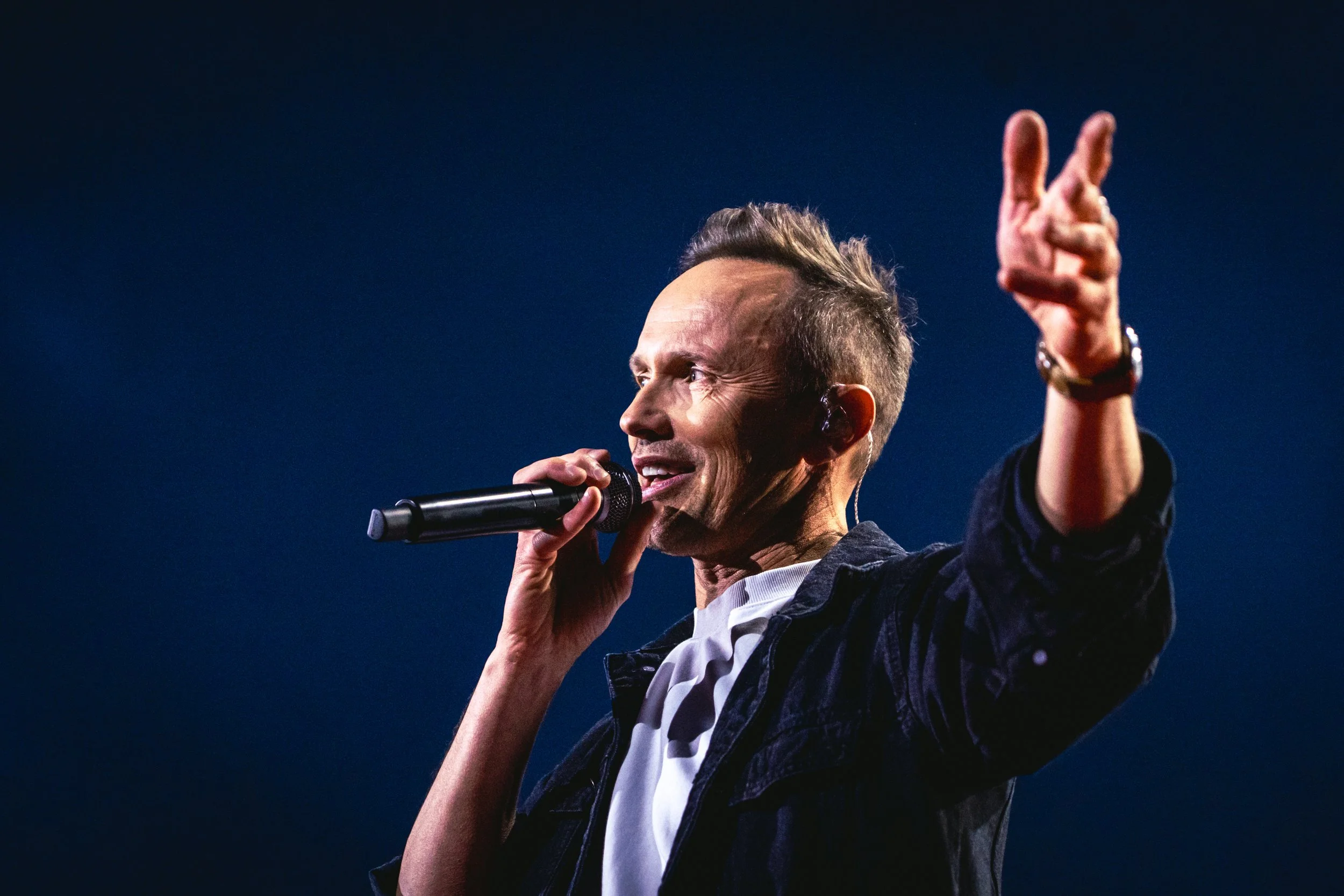 A man singing into a microphone on stage, making a peace sign with his right hand, against a dark blue background. Prata Vetra. Renars Kaupers