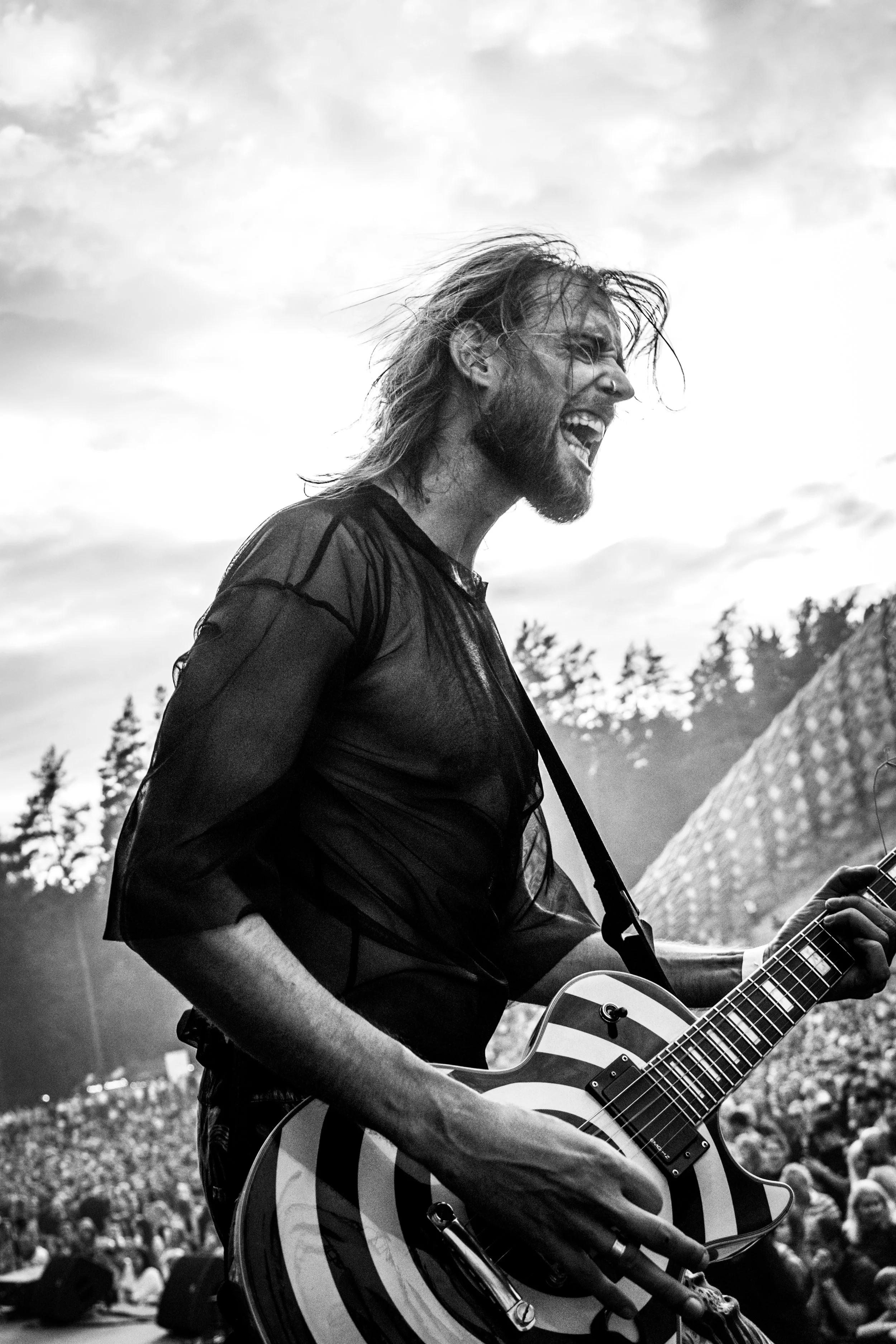 A man with long hair and a beard is smiling widely and playing an acoustic guitar. He is outdoors with a cloudy sky and trees in the background, and a crowd is gathered around him.