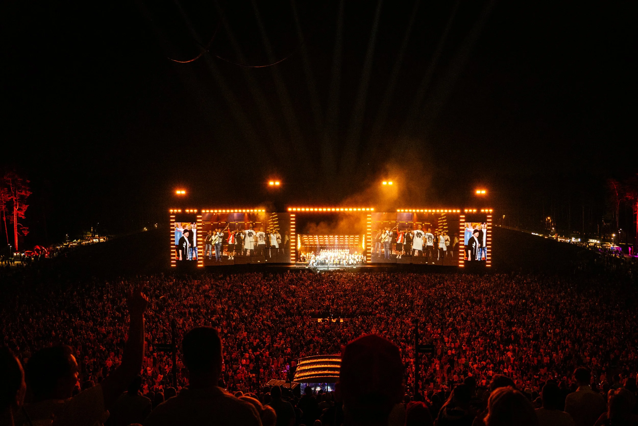 A live outdoor concert at night with a large crowd, brightly lit stage with performer images and musicians, and bright lighting above.