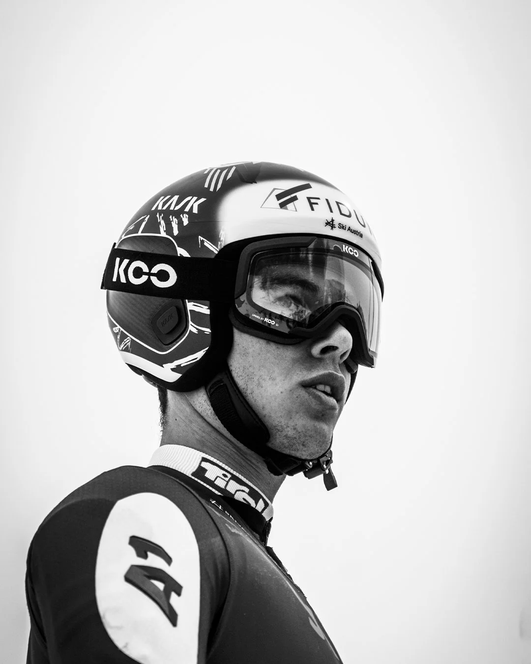 A black and white photo of a skier wearing a helmet and goggles, looking to the side.