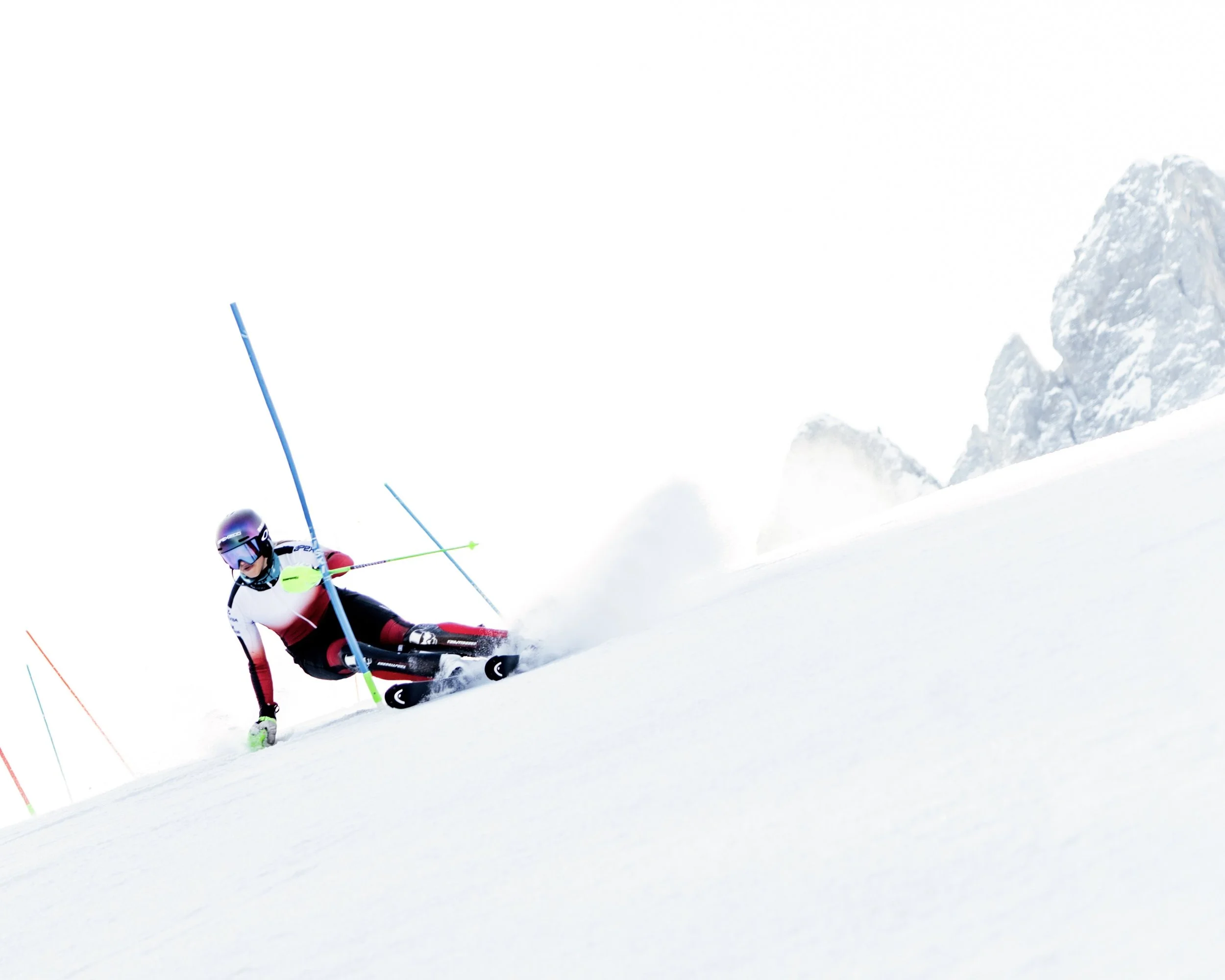 A skier in a white, red, and black suit skiing downhill on snow. The skier is wearing a helmet and goggles, and is leaning into a turn with ski poles. Snow-covered mountain peaks are visible in the background.