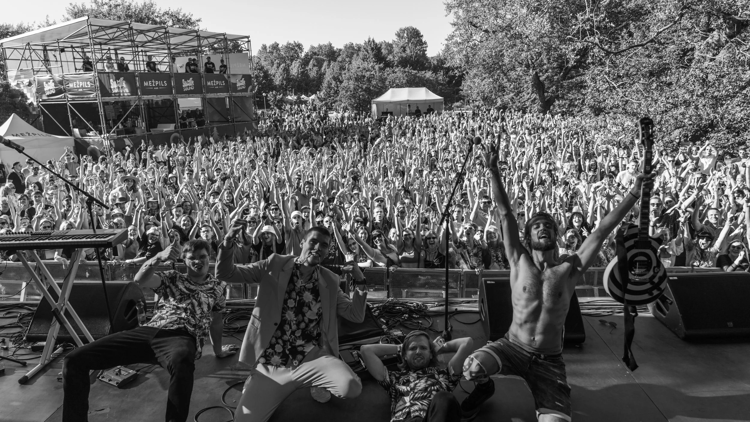 A large outdoor music festival with a stage and a crowd of excited attendees, some sitting and some standing on the stage with musical instruments, during daytime.
