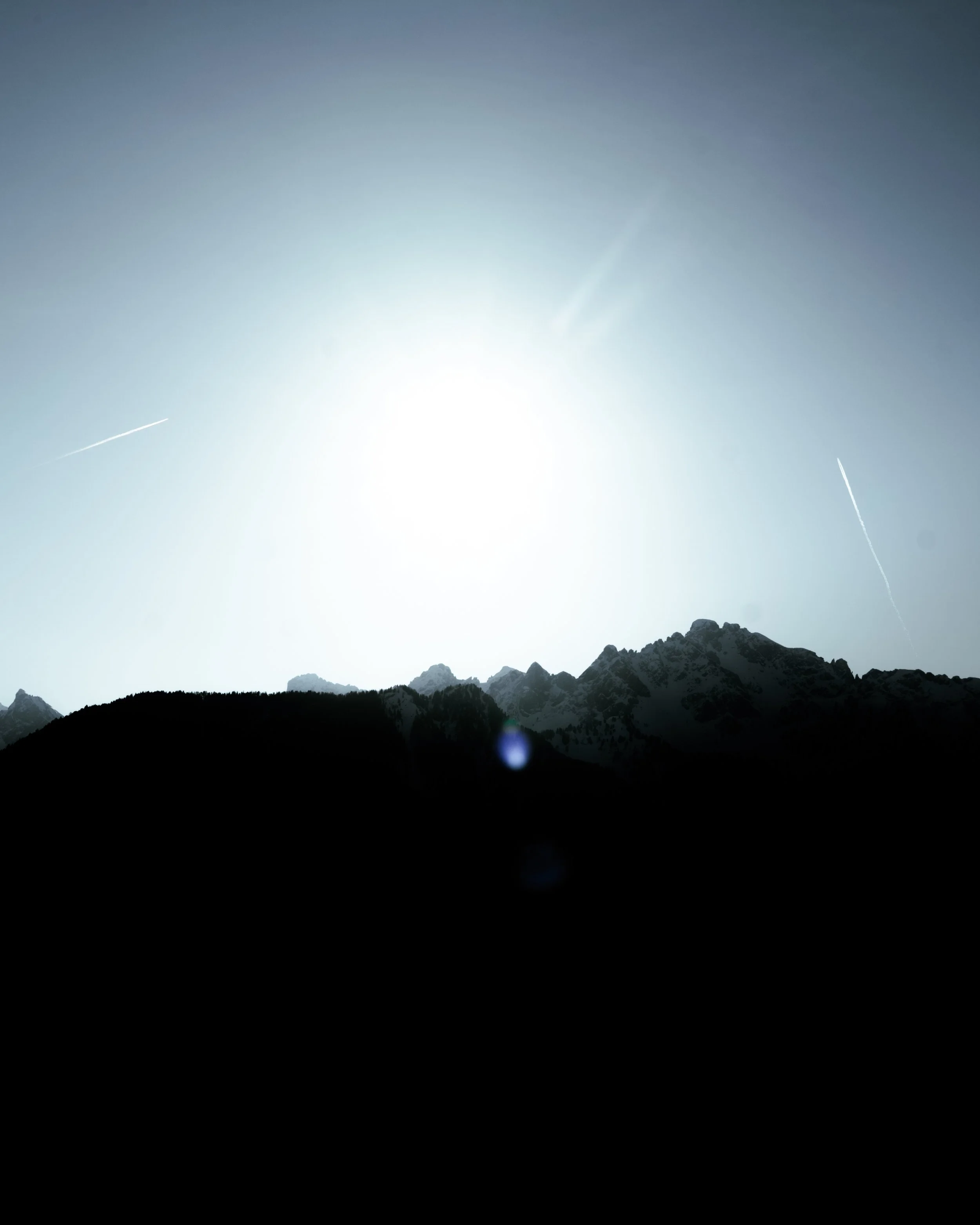 Snow-capped mountain peaks under a bright sun in a clear sky with contrails visible above.