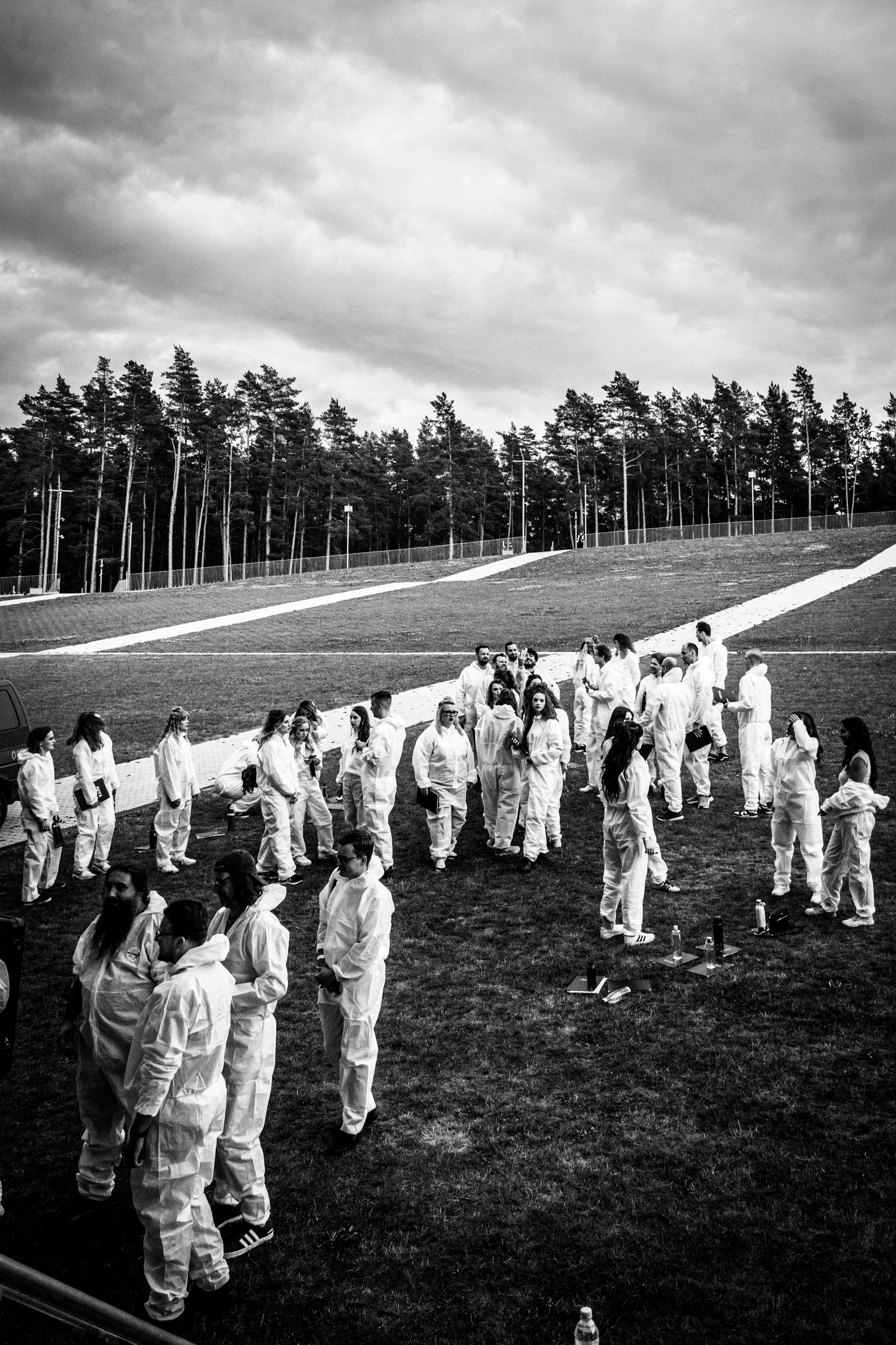 A group of people dressed in white uniforms standing outside on grass, preparing for a martial arts class or event, with a backdrop of tall trees and a cloudy sky.