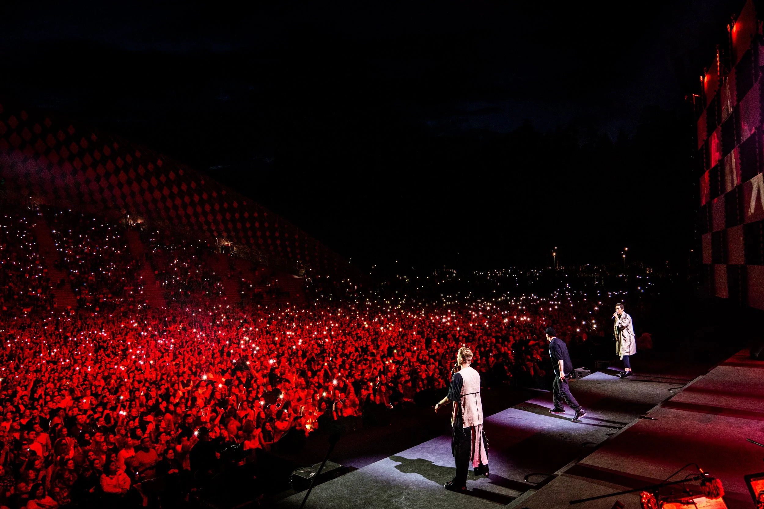 Concert stage with performers and a large audience holding up lights, night sky visible in the background. Lielkoncerts