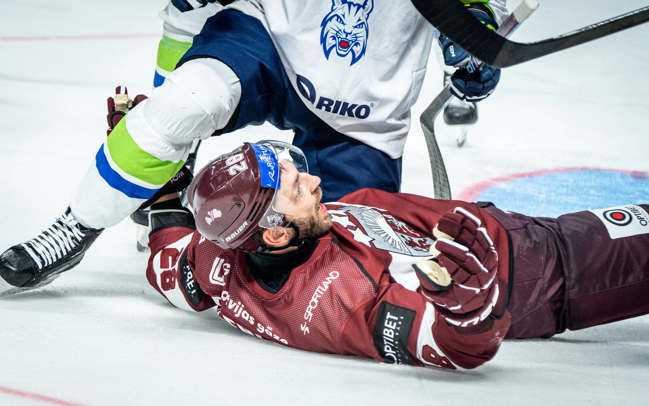 Hockey players on the ice, one player in maroon lying on the ice with a beard and helmet, another player in a white and blue jersey kneeling beside him, appearing to check or help him after a fall during a game.