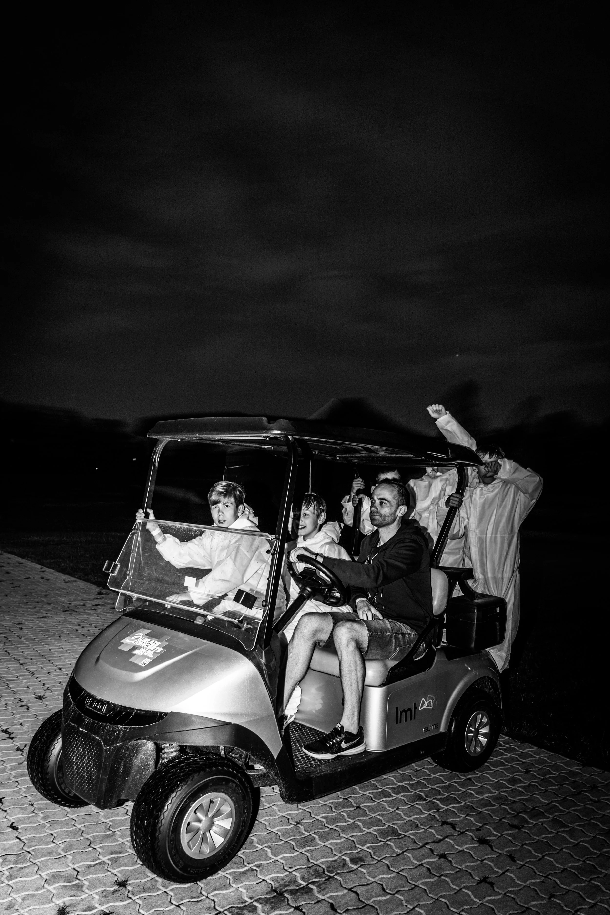 A group of children and adults riding in a golf cart at night. The kids are wearing jackets, and one child is waving with a raised fist. The golf cart is on a paved surface, and the sky is dark with some clouds visible. Lielkoncerts. Backstage