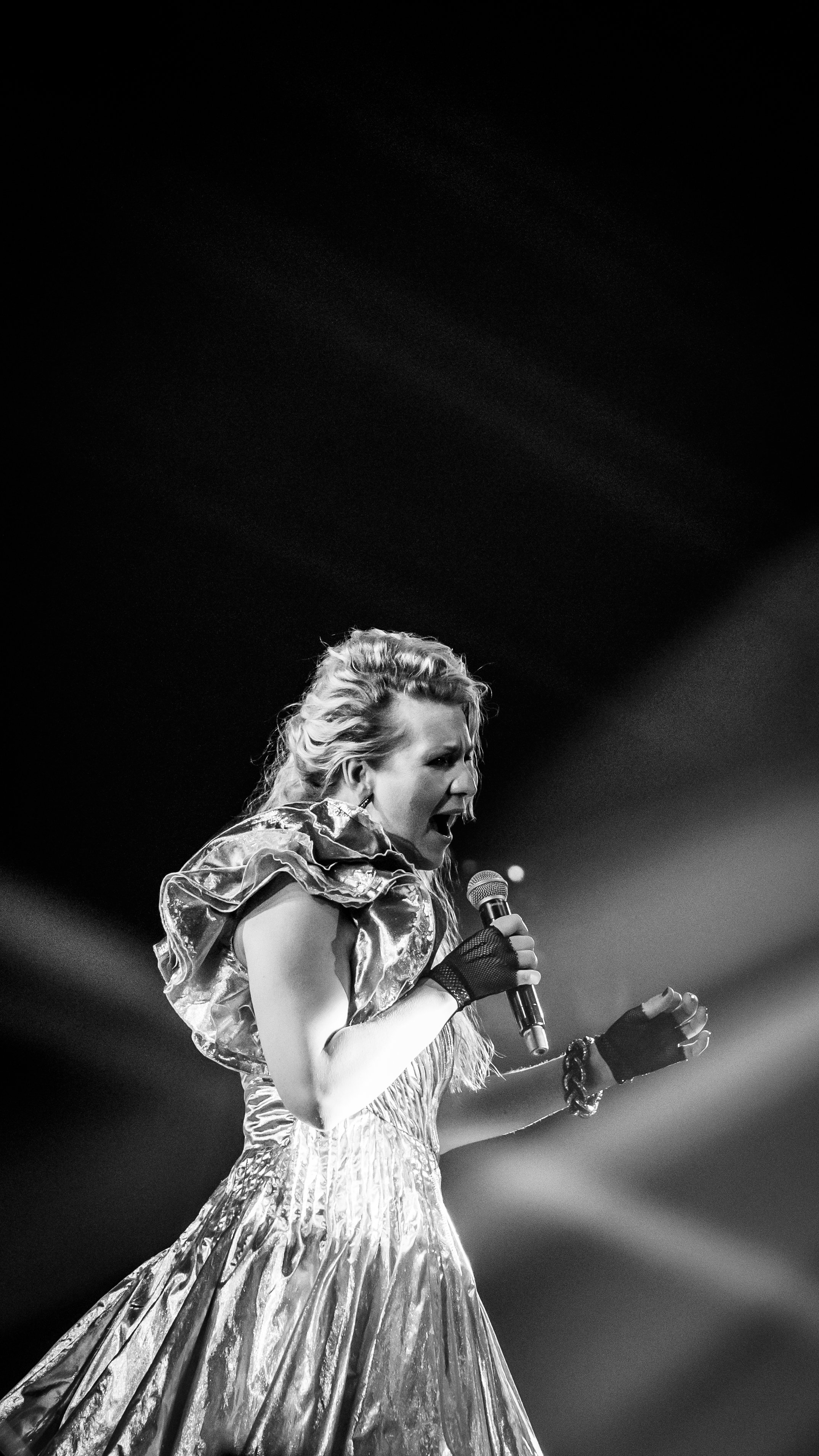 Black and white photo of a woman singing passionately into a microphone on stage, wearing an elaborate, shiny dress with ruffled sleeves.