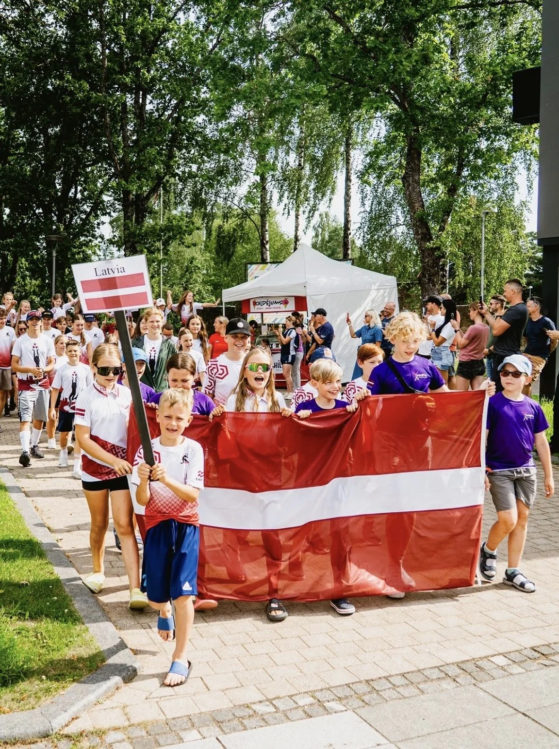 Children participating in a parade holding a Latvian flag, with spectators and trees in the background.