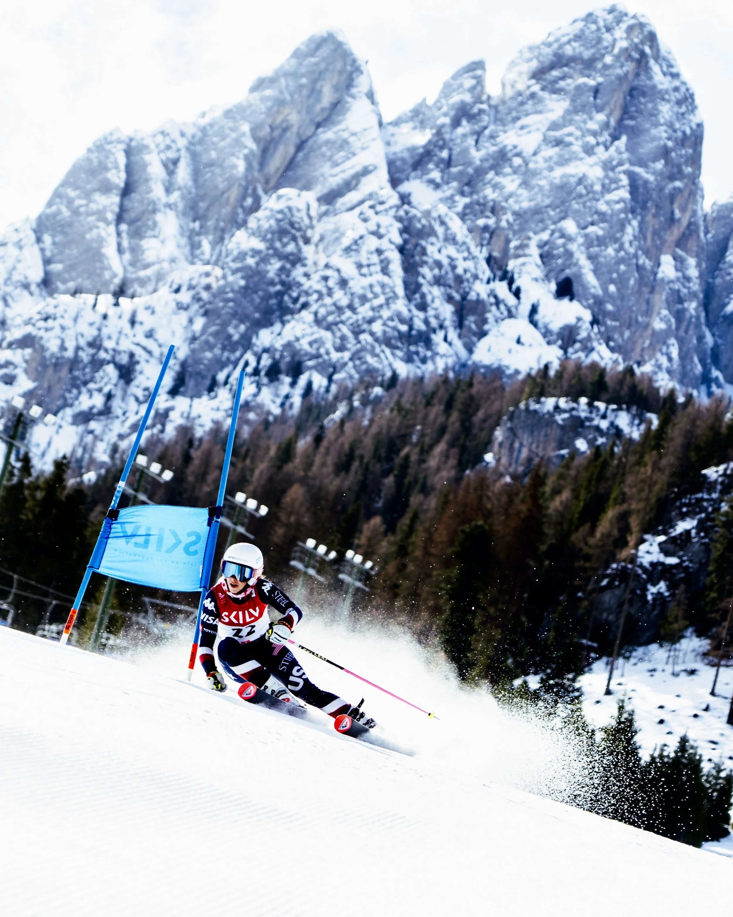 A skier in a red, black, and white uniform racing downhill on snow-covered slopes with mountains in the background.