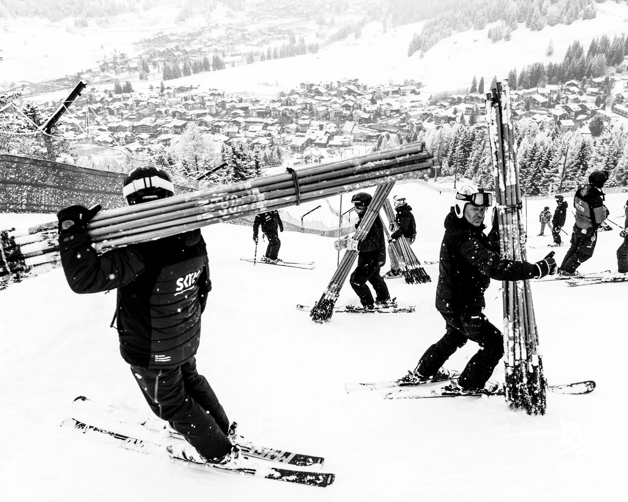 People skiing down a snowy slope with a town and snow-covered trees in the background.