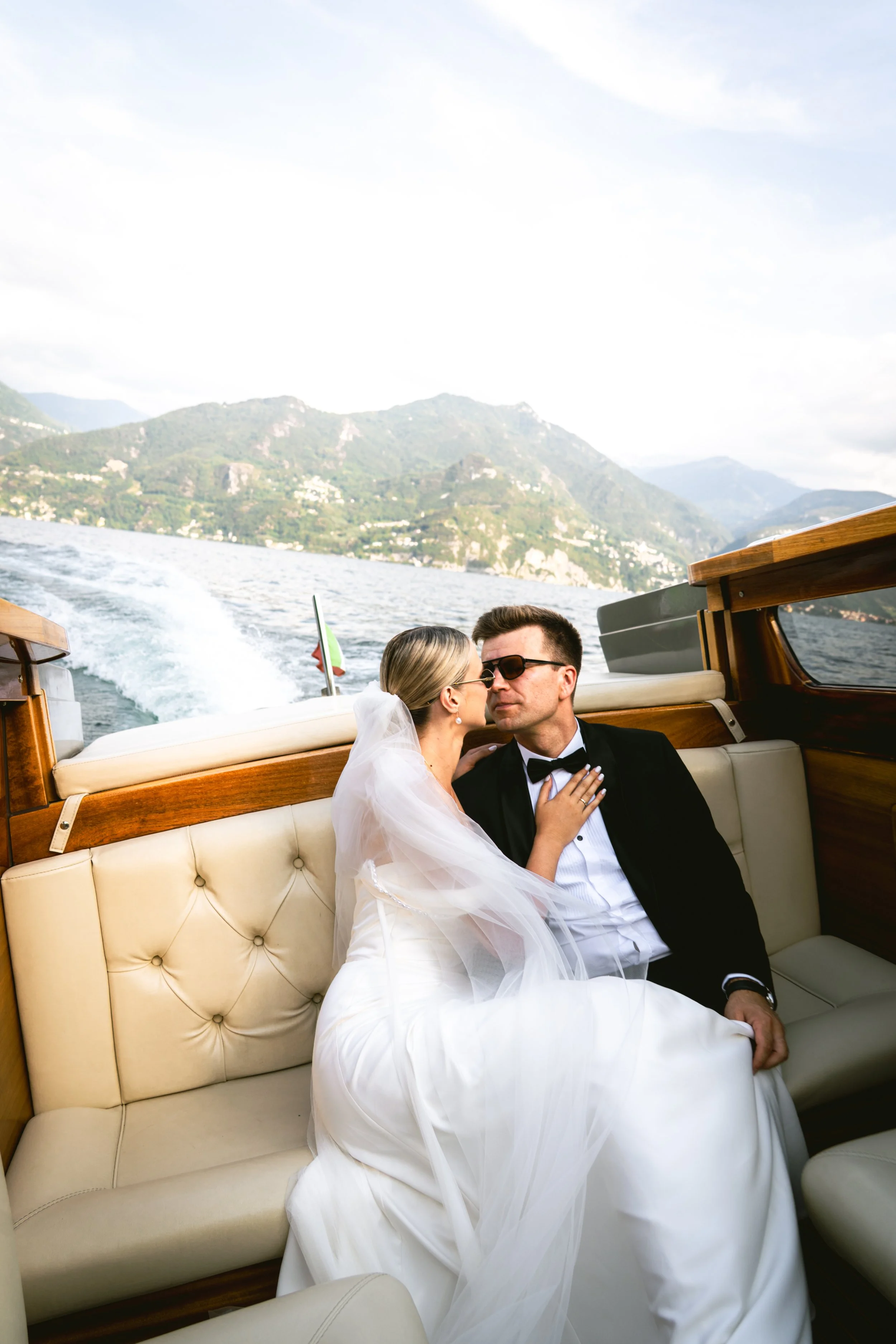 A bride and groom sitting closely on a boat with a scenic lake and mountains in the background, celebrating their wedding.