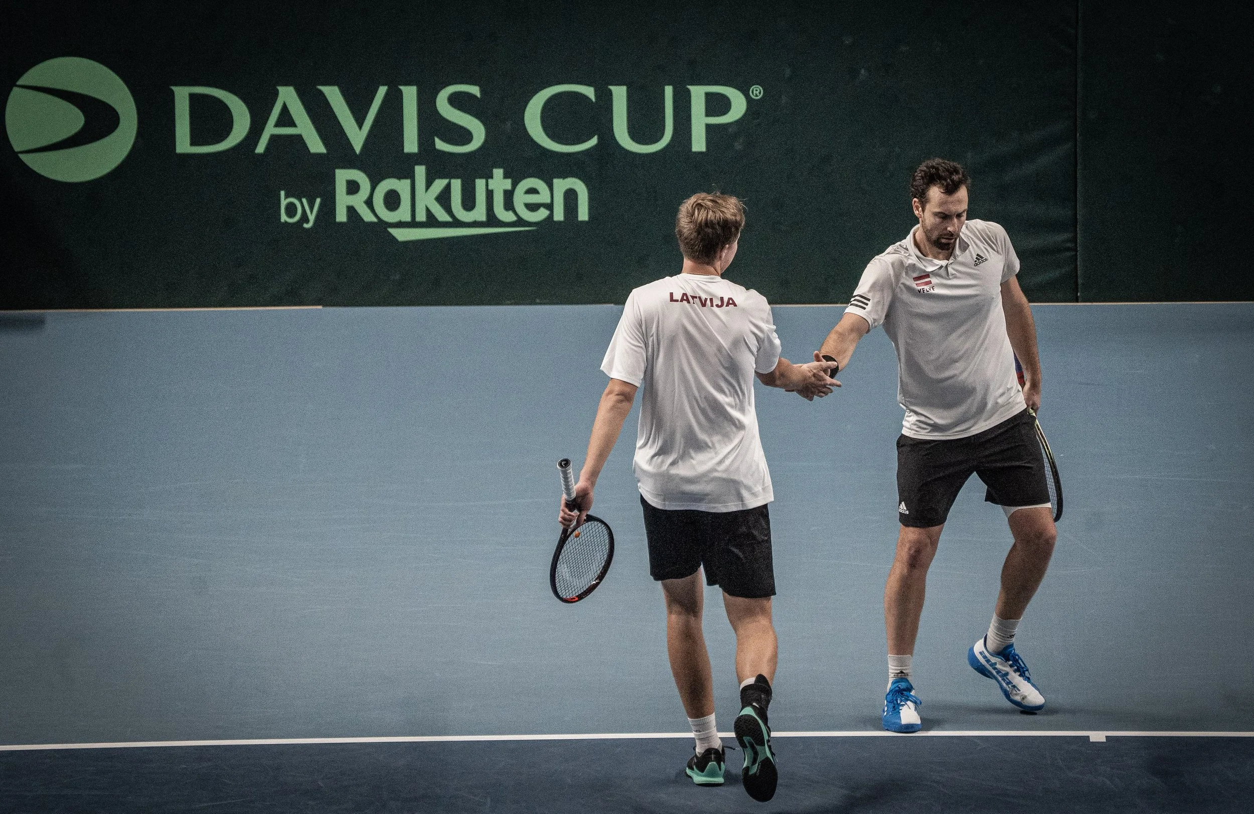Two male tennis players on an indoor court, one with his back to the camera and wearing a white shirt with 'LARVIJA' on the back, and the other facing forward, both holding tennis rackets. A large green sign with the text 'Davis Cup by Rakuten' is vi