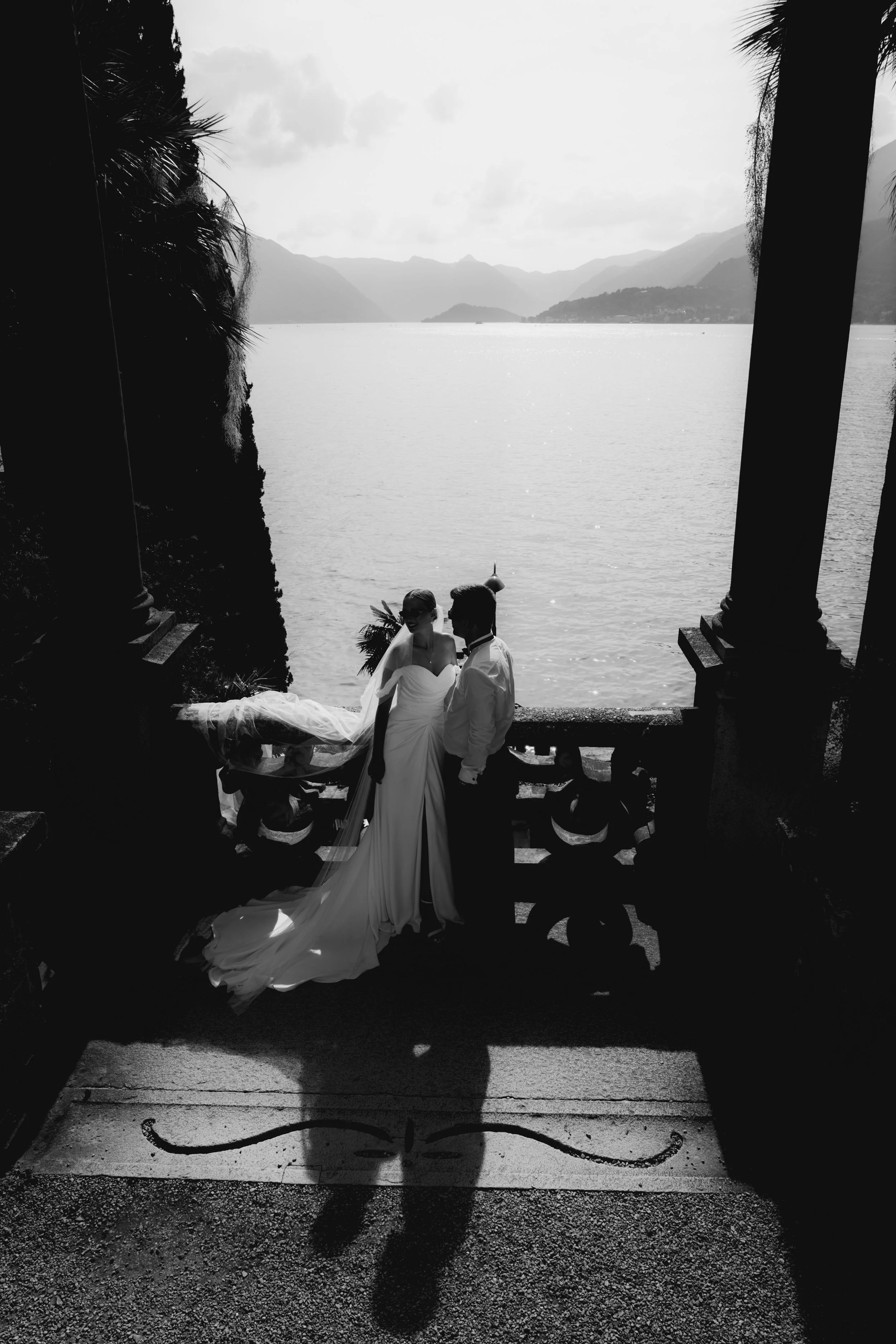 A black and white photo of a bride and groom standing on a staircase outdoors, with a lake and mountainous landscape in the background. The bride is wearing a strapless wedding gown and veil, and the groom is in a suit. Shadows of trees and the coupl