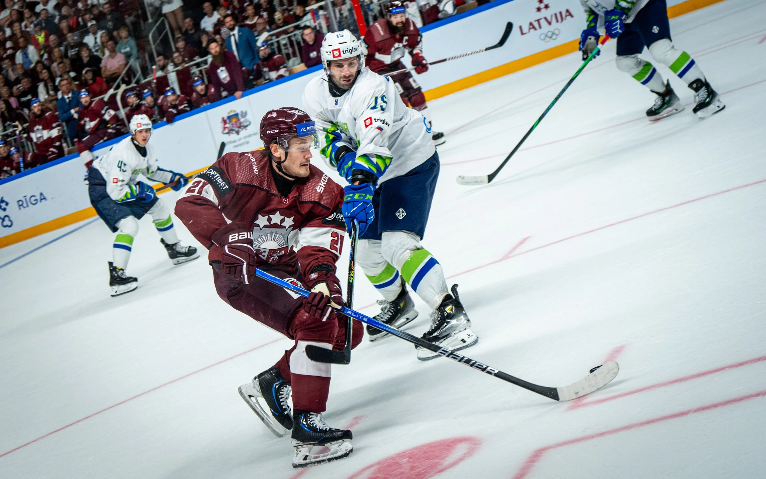 Two ice hockey players are engaged in ongoing play on the rink, with a crowd watching from the stands. One player in a maroon jersey is skating forward, while the other in a white jersey appears to be battling for control of the puck.