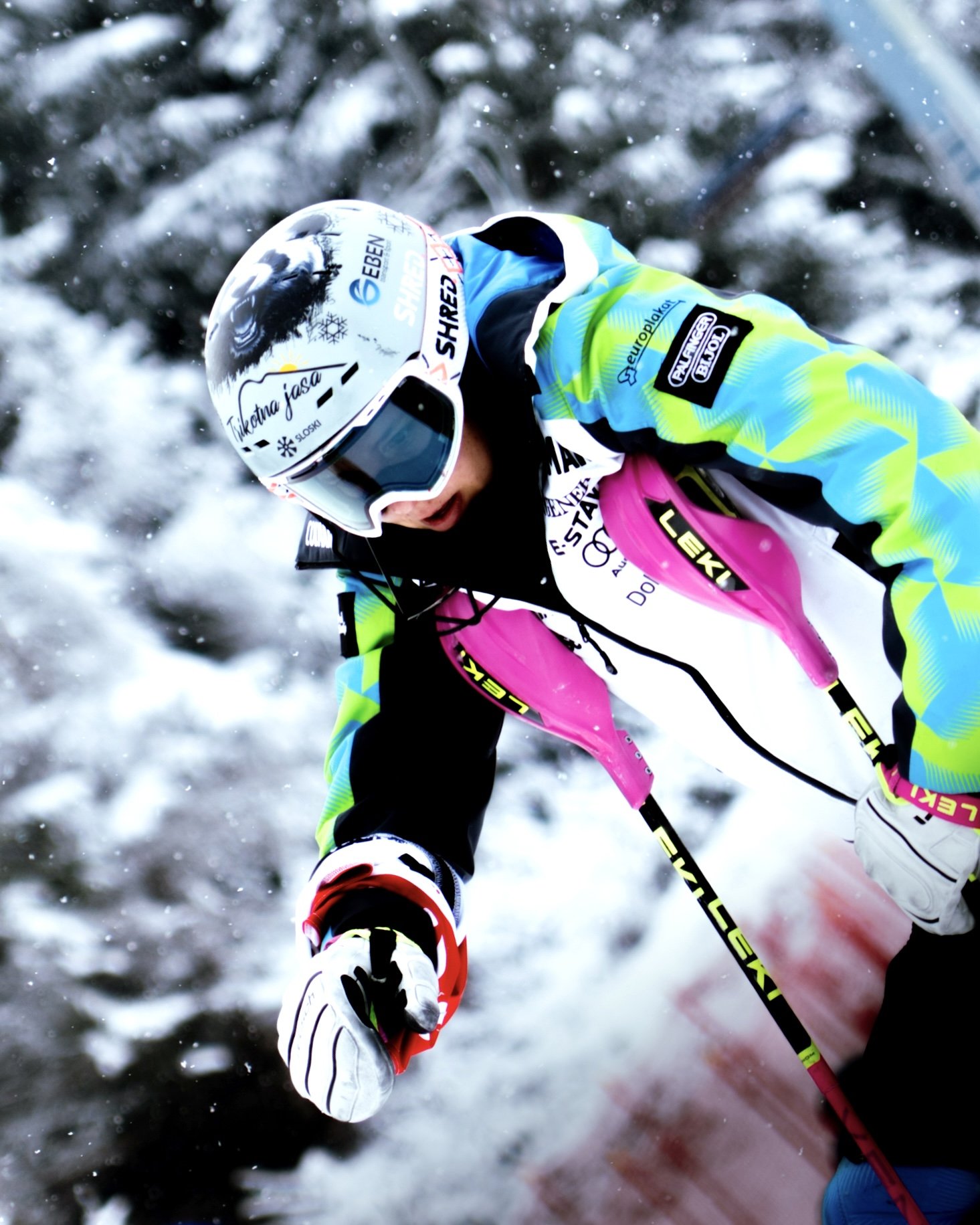 A cross-country skier wearing a helmet, goggles, and bright sports gear actively skiing through snow, with trees and snow-covered scenery in the background.