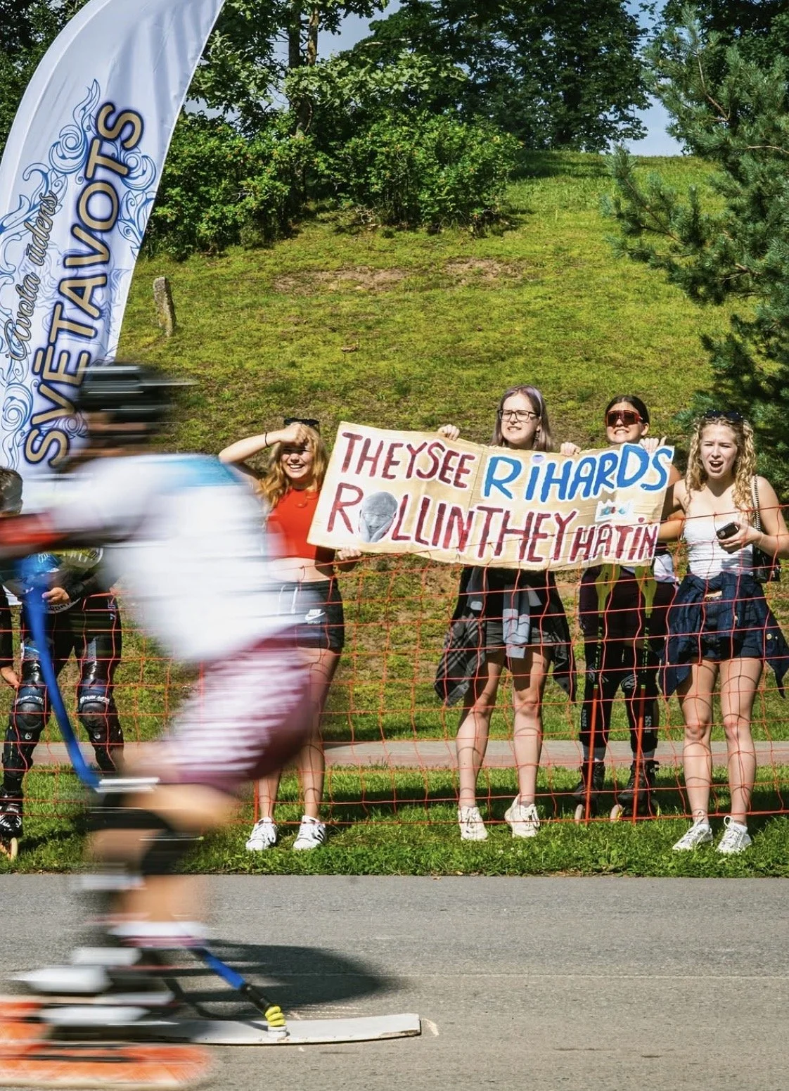 Group of young women holding a protest sign behind a temporary orange fence near a skateboarding area, with a blurred skateboarder passing by in the foreground. The sign reads, "They see Richard rollin' in the Hatin'." There is a banner on the left s